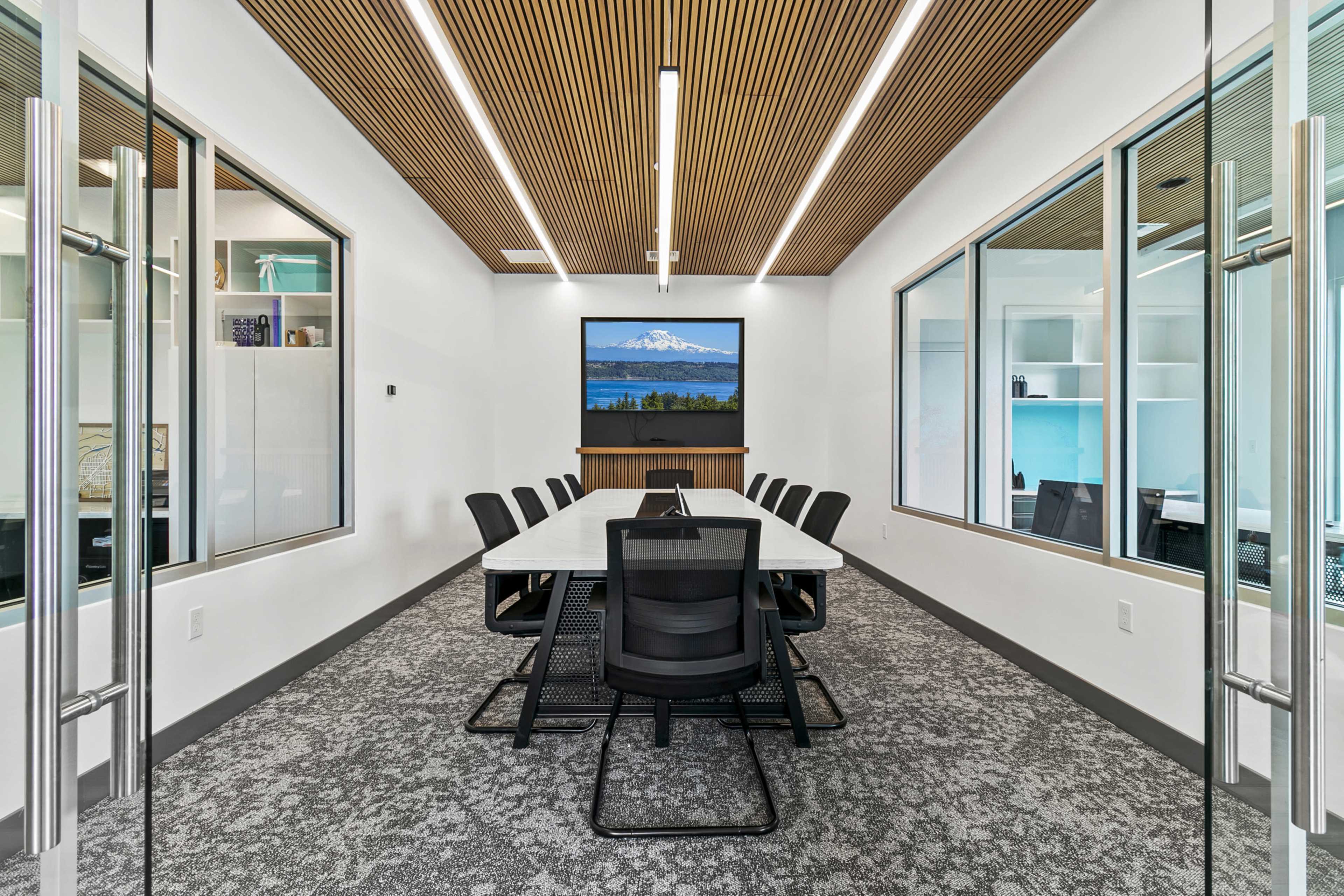 A modern conference room features a long table surrounded by black chairs, with a large screen displaying a mountain landscape in the background and glass walls allowing for visibility from the hallway.