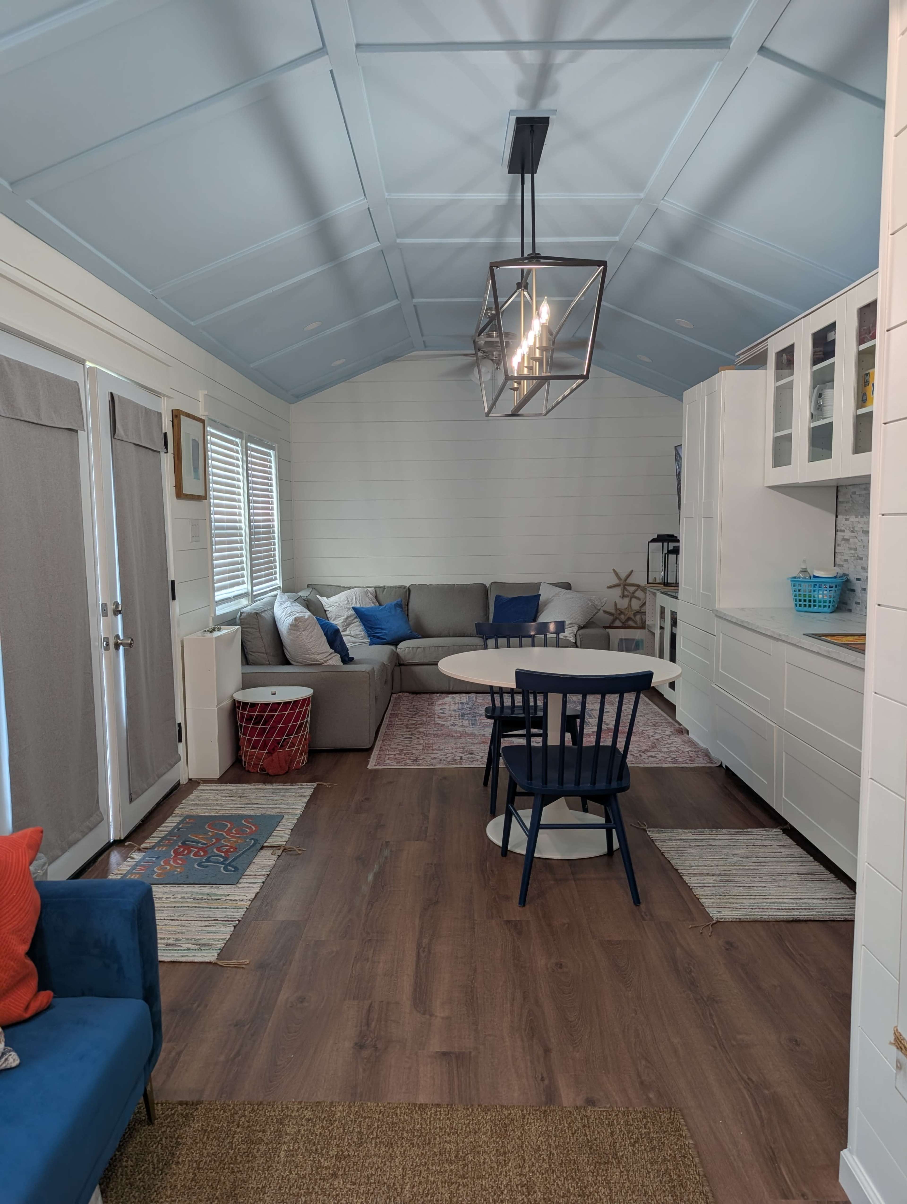 The image shows the interior of a small, modern cabin featuring a living area with a sectional sofa, a dining table with chairs, and a kitchen area with white cabinetry.