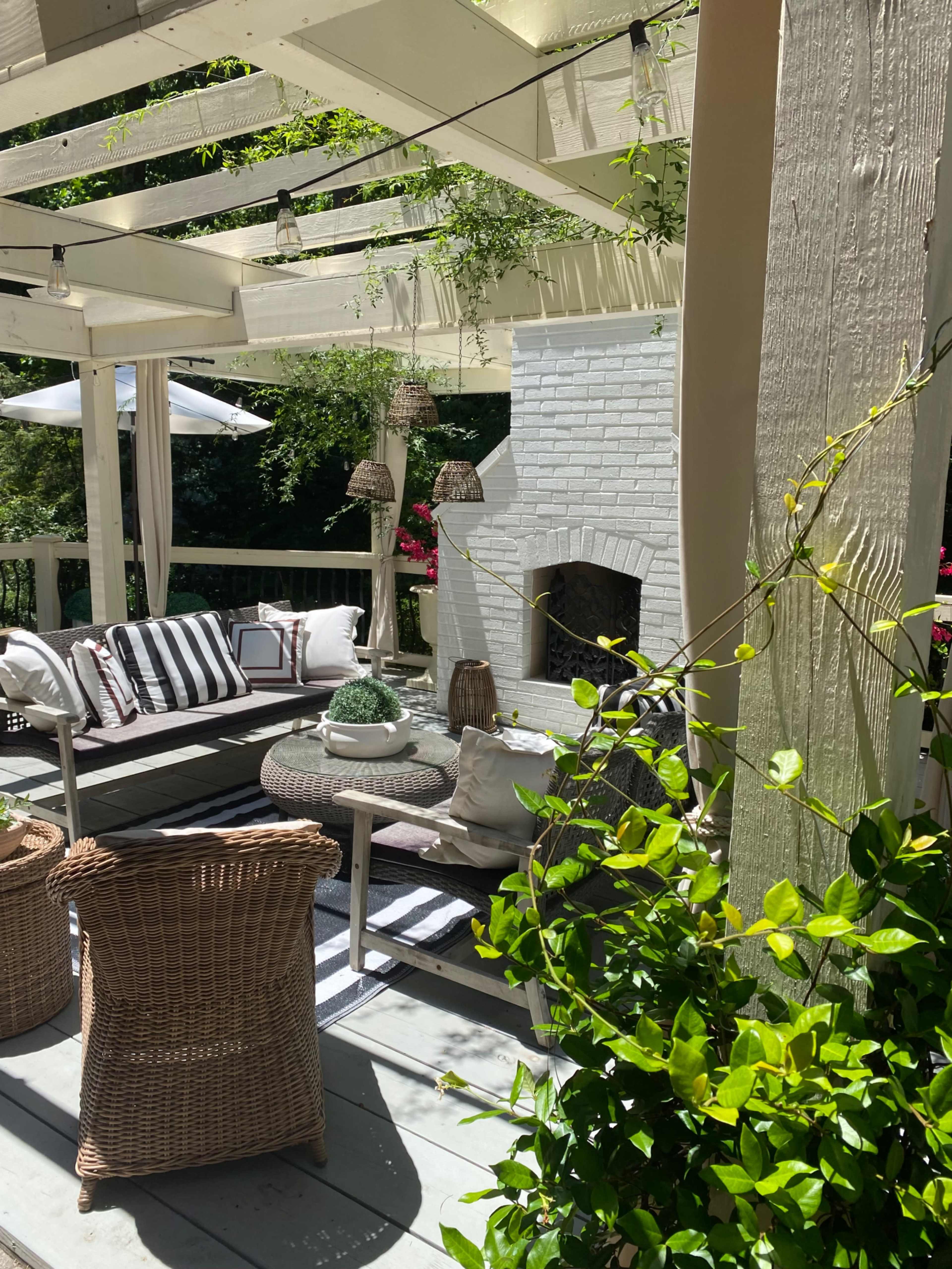 A cozy outdoor seating area features a white brick fireplace, woven furniture, and potted plants under a pergola.