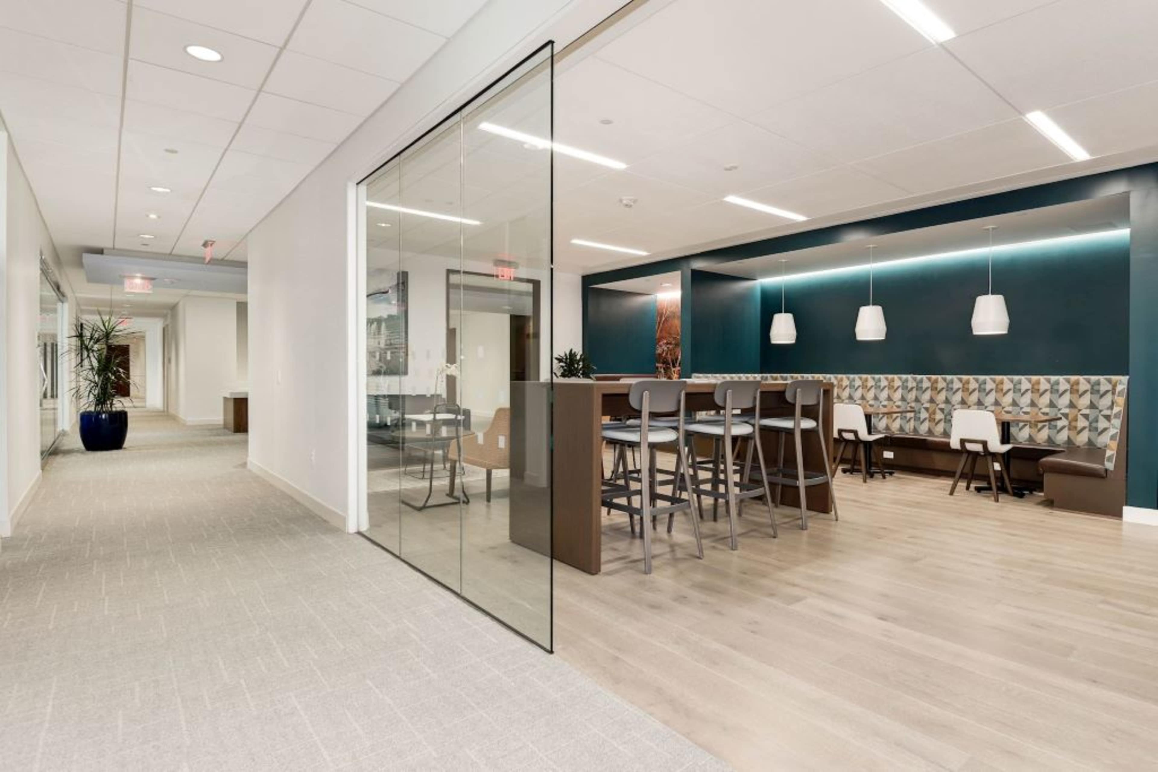 A modern office space with a glass partition separating a communal seating area featuring high tables and chairs from a corridor lined with plants.