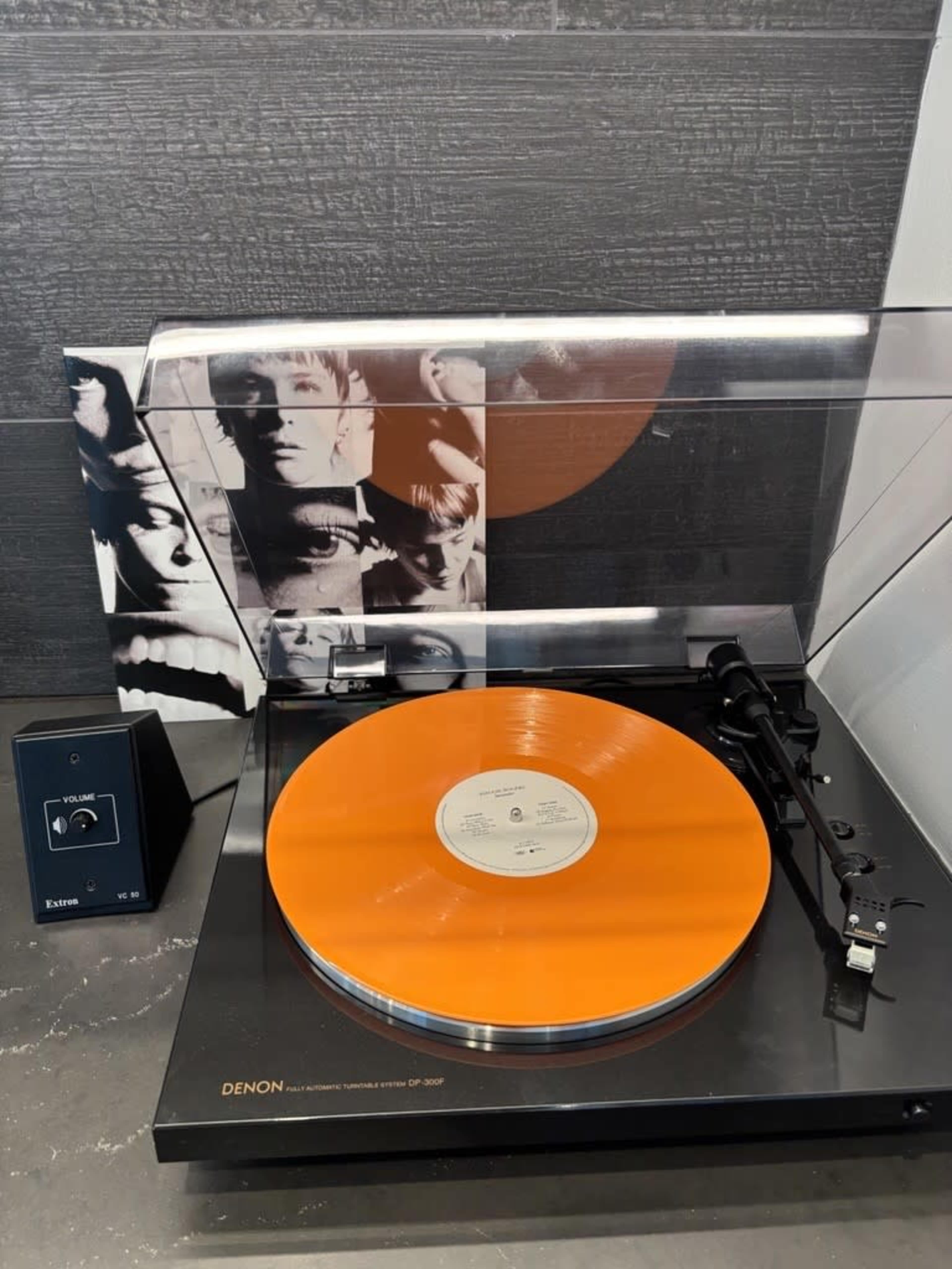 A bright orange vinyl record is placed on a black turntable, accompanied by a cover featuring black and white images of faces.