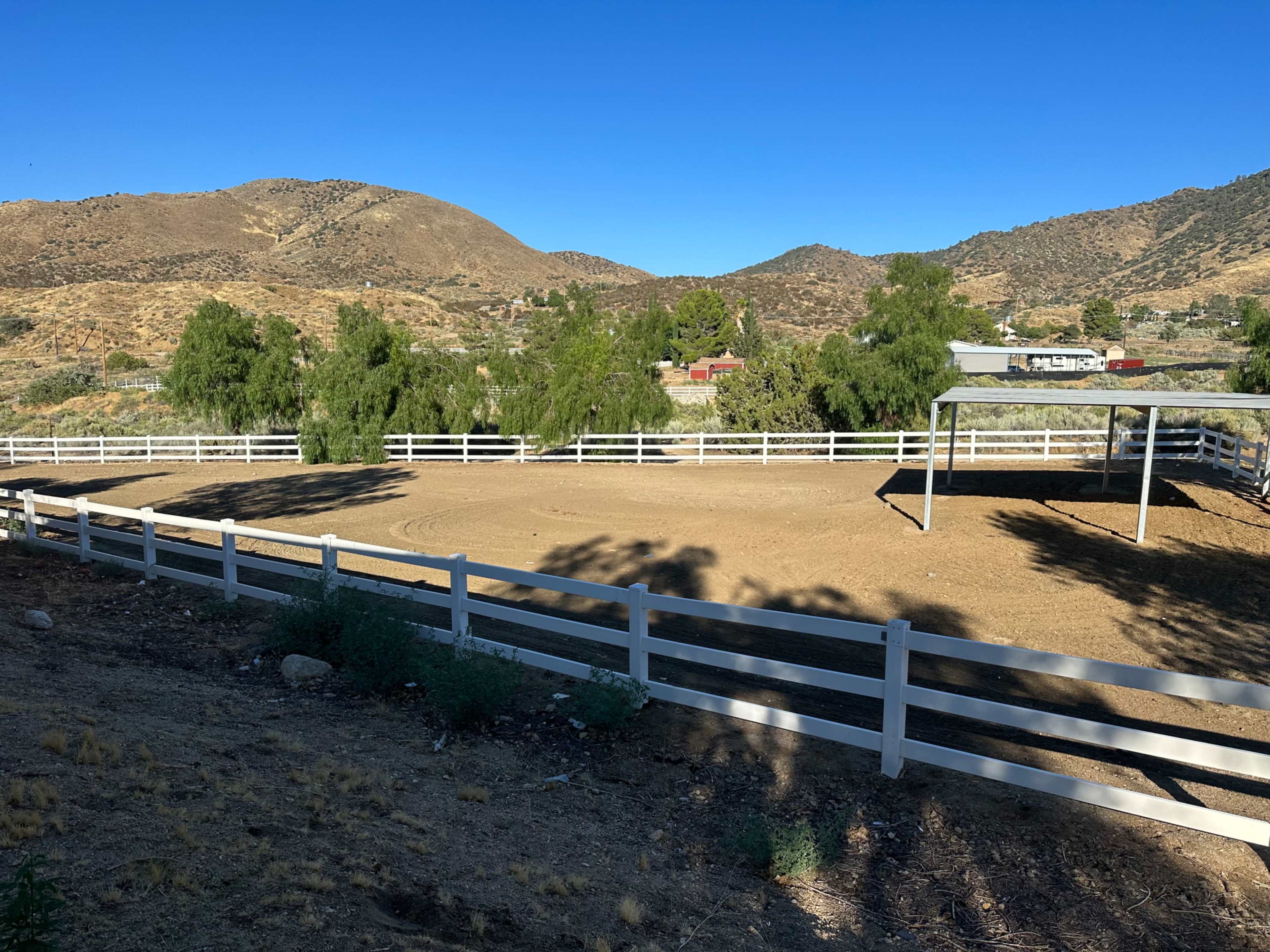 A horse riding arena surrounded by white fencing, set against a backdrop of rolling hills and clear blue skies.