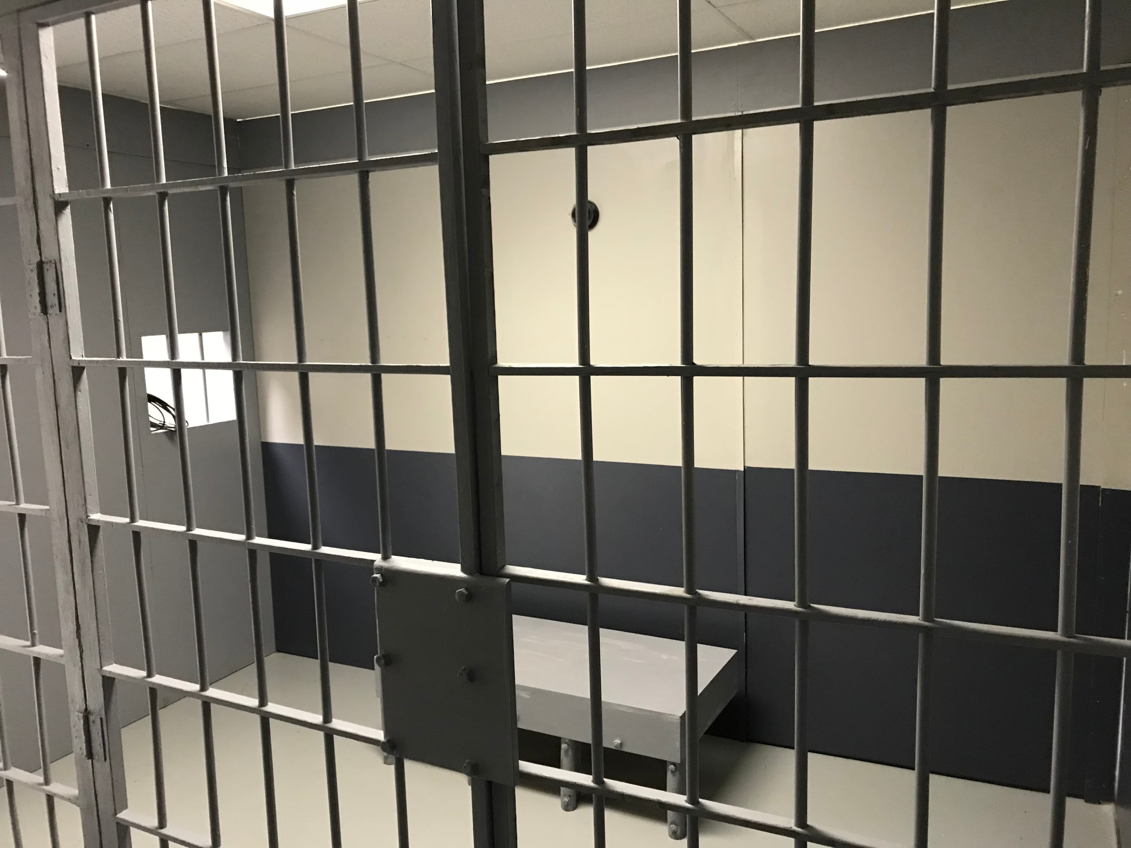 The image shows the inside of a small, empty jail cell with bars and a plain concrete table.