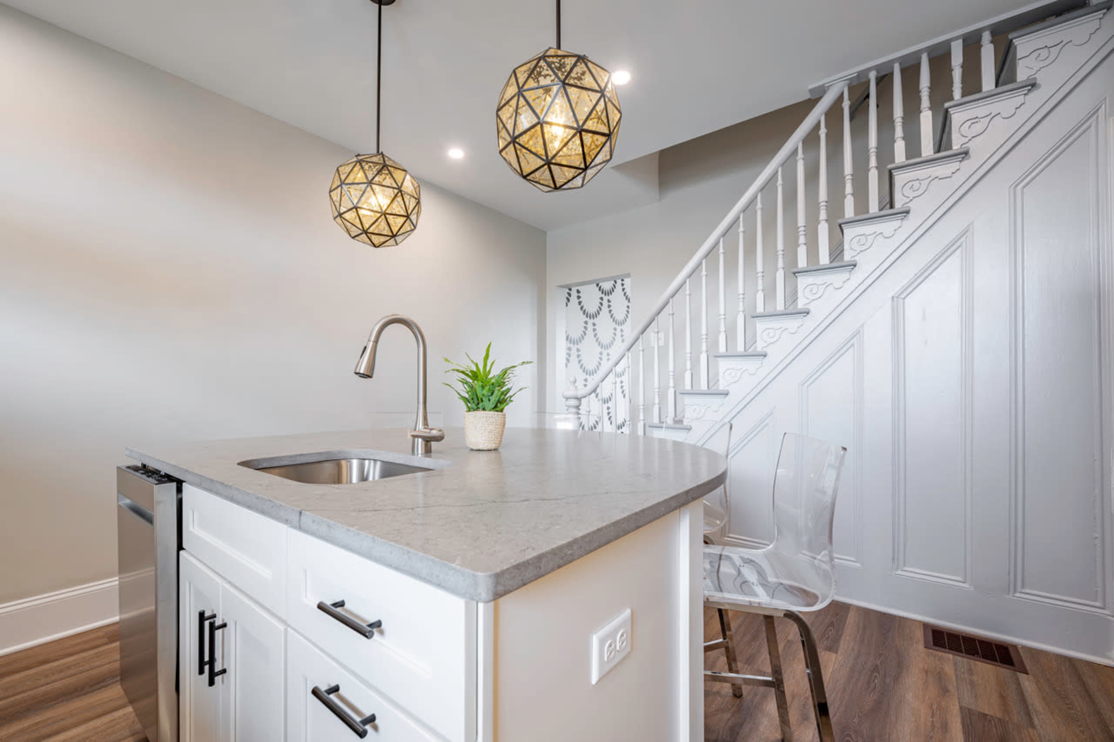 The image shows a modern kitchen area with a concrete countertop, a stainless steel sink, and two geometric pendant lights, adjacent to a staircase.