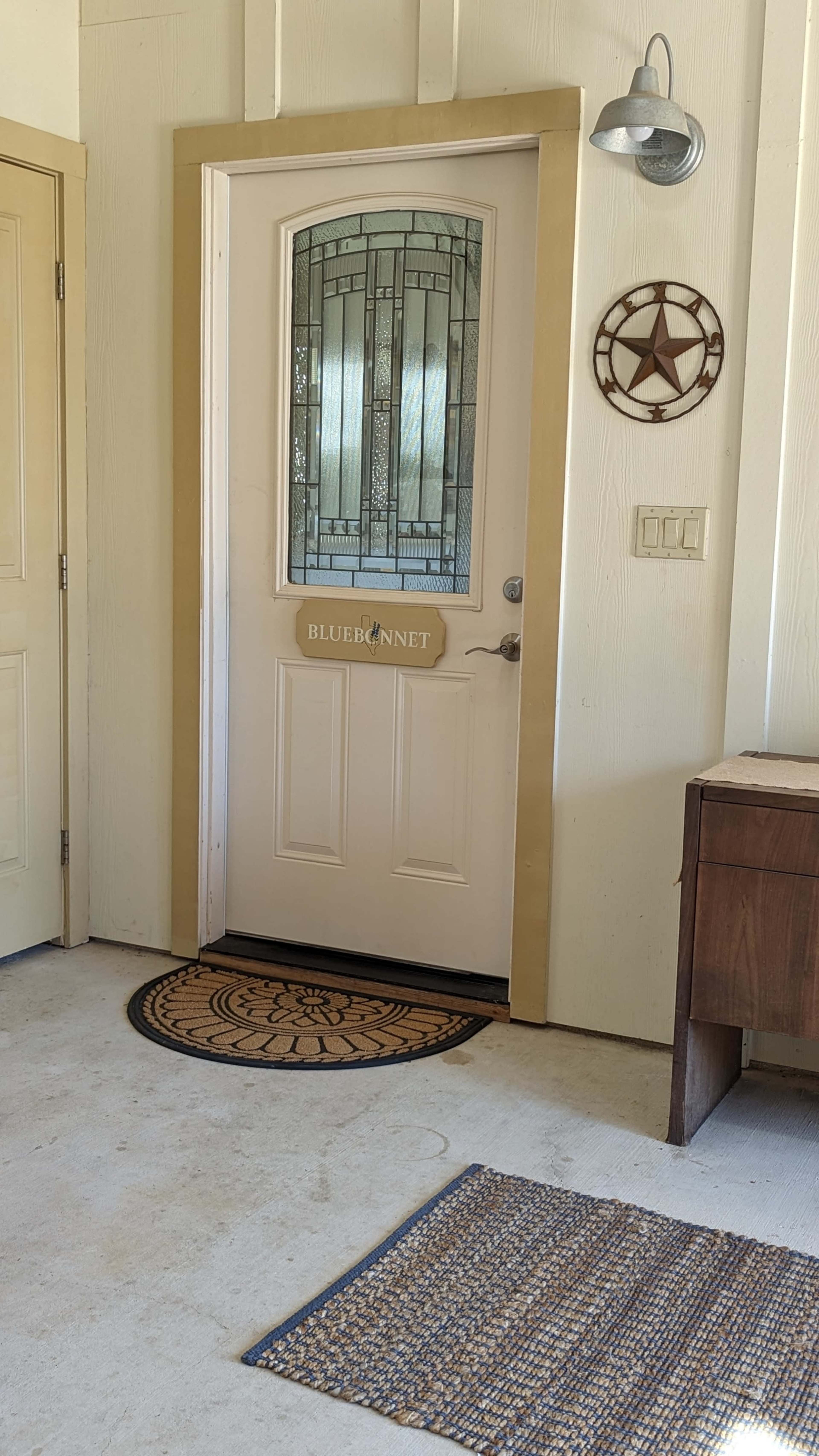 A front door with decorative glass, featuring a small sign labeled "BLUEBONNET," alongside a star-shaped wall decoration and two rugs on the floor.