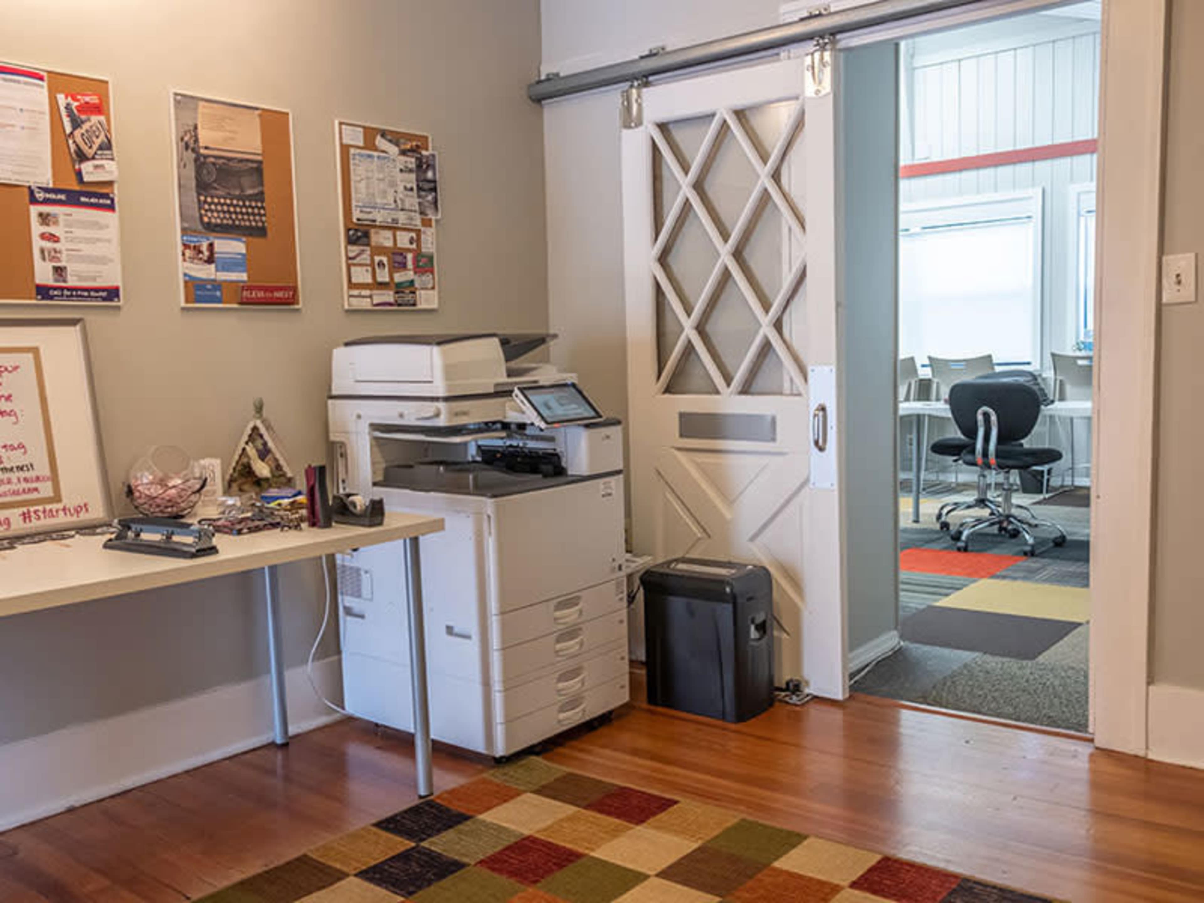 An office space featuring a printer beside a table with various notices and a doorway leading to a room with chairs and carpet.