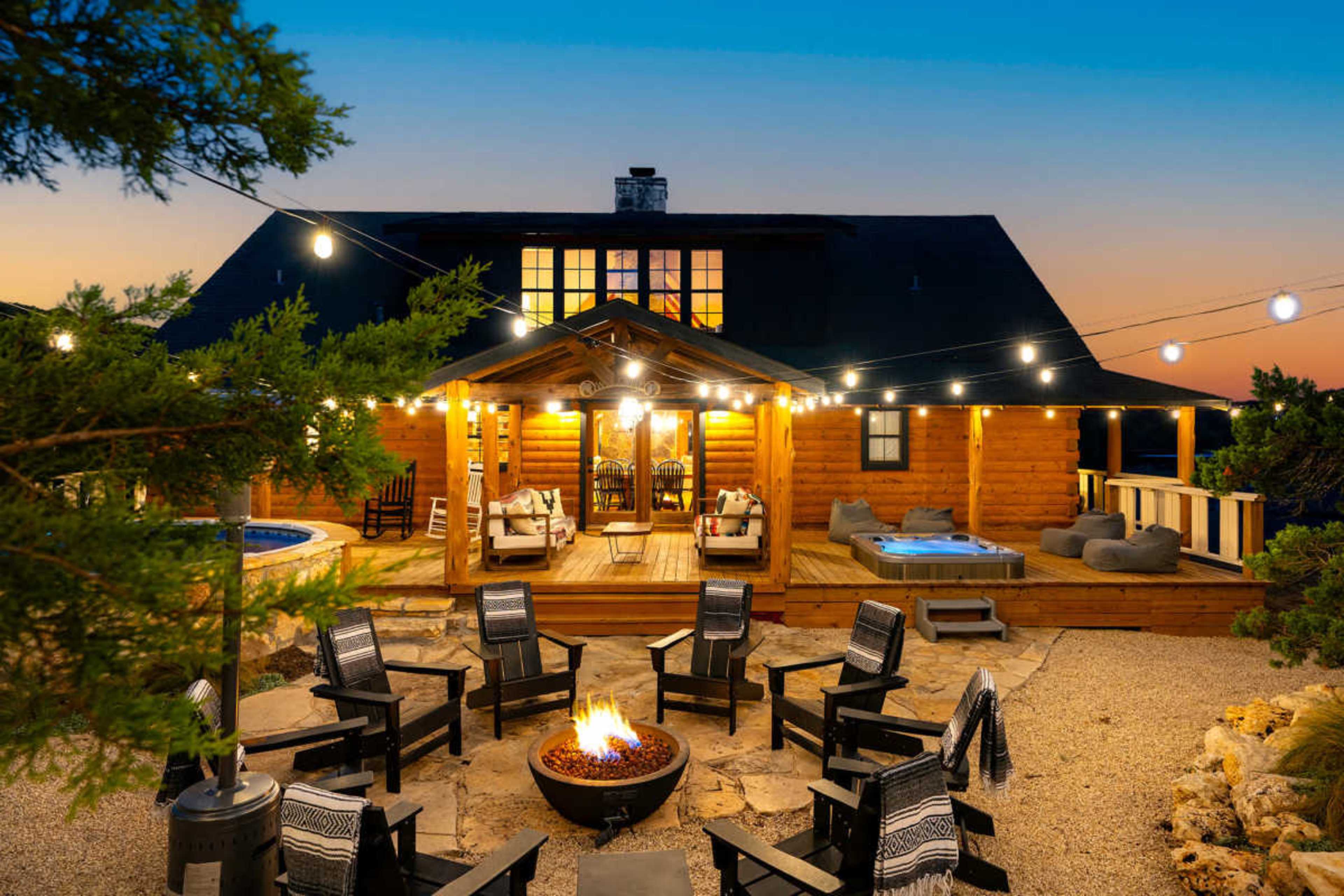 A log cabin with a large outdoor seating area and fire pit is illuminated by string lights at dusk.