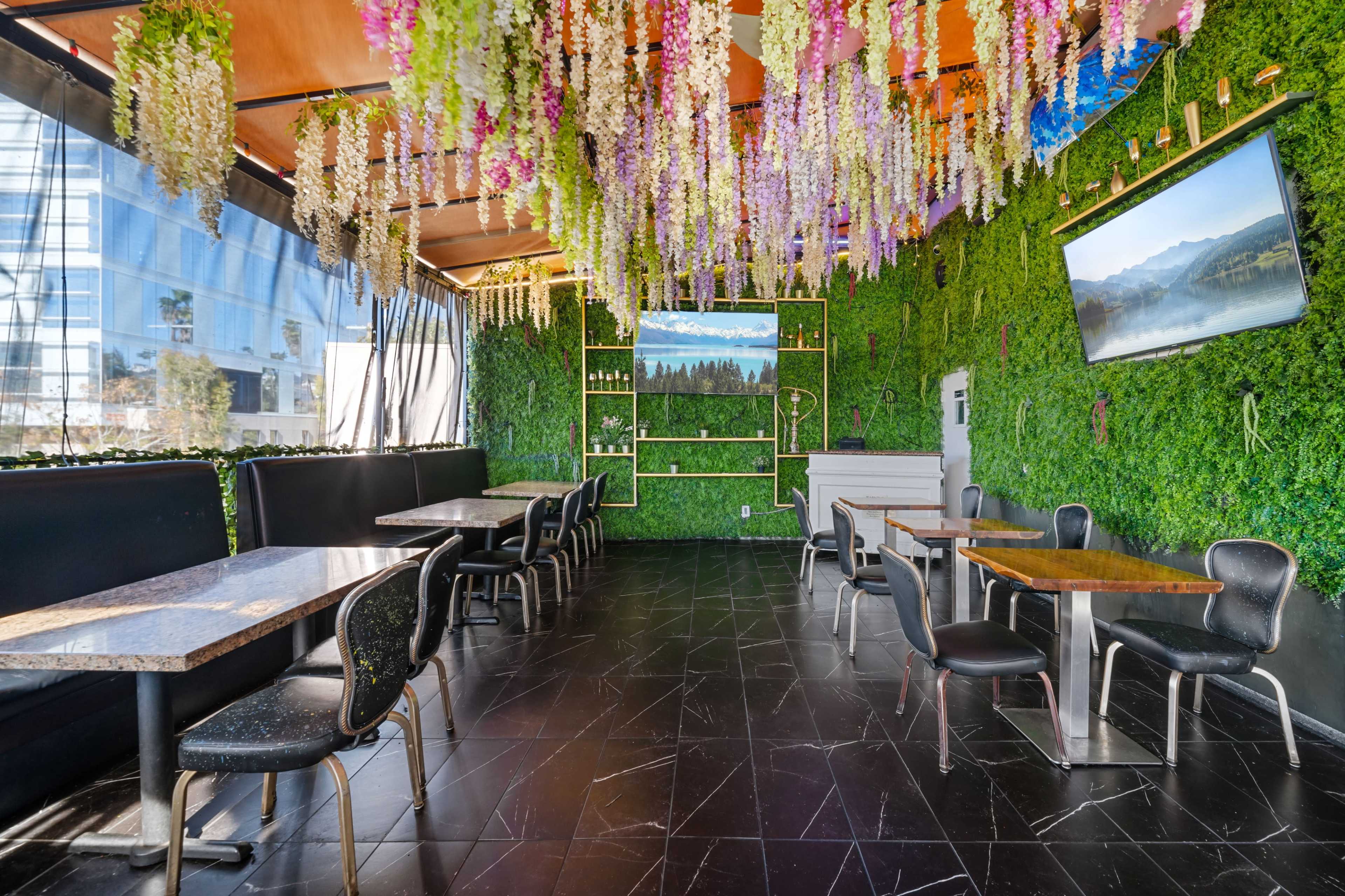 The image shows an indoor restaurant with green wall decor, tables, and chairs, set beneath hanging floral elements.