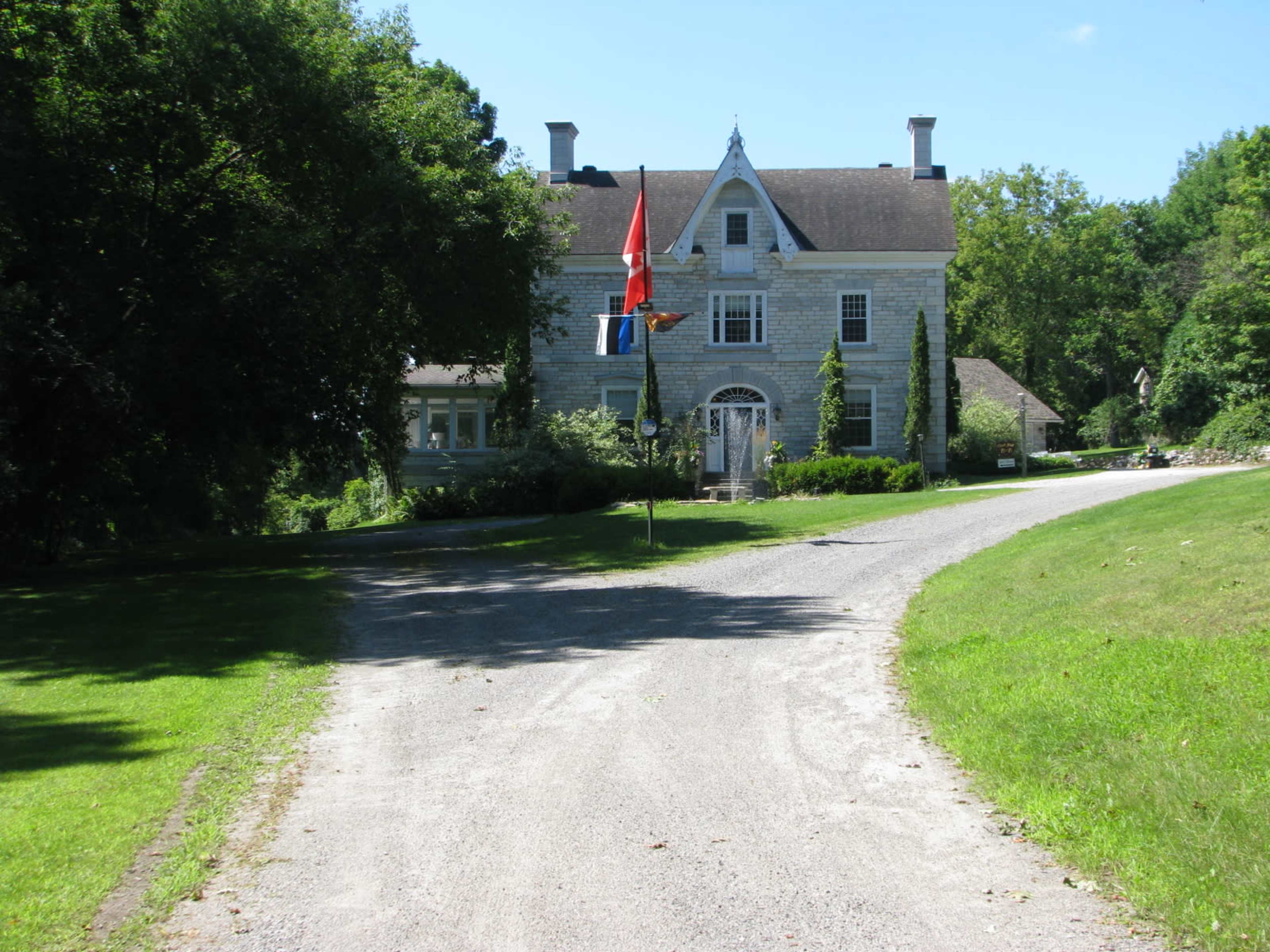 A gravel driveway leads up to a large, historic house surrounded by trees and greenery, with flags displayed in front.