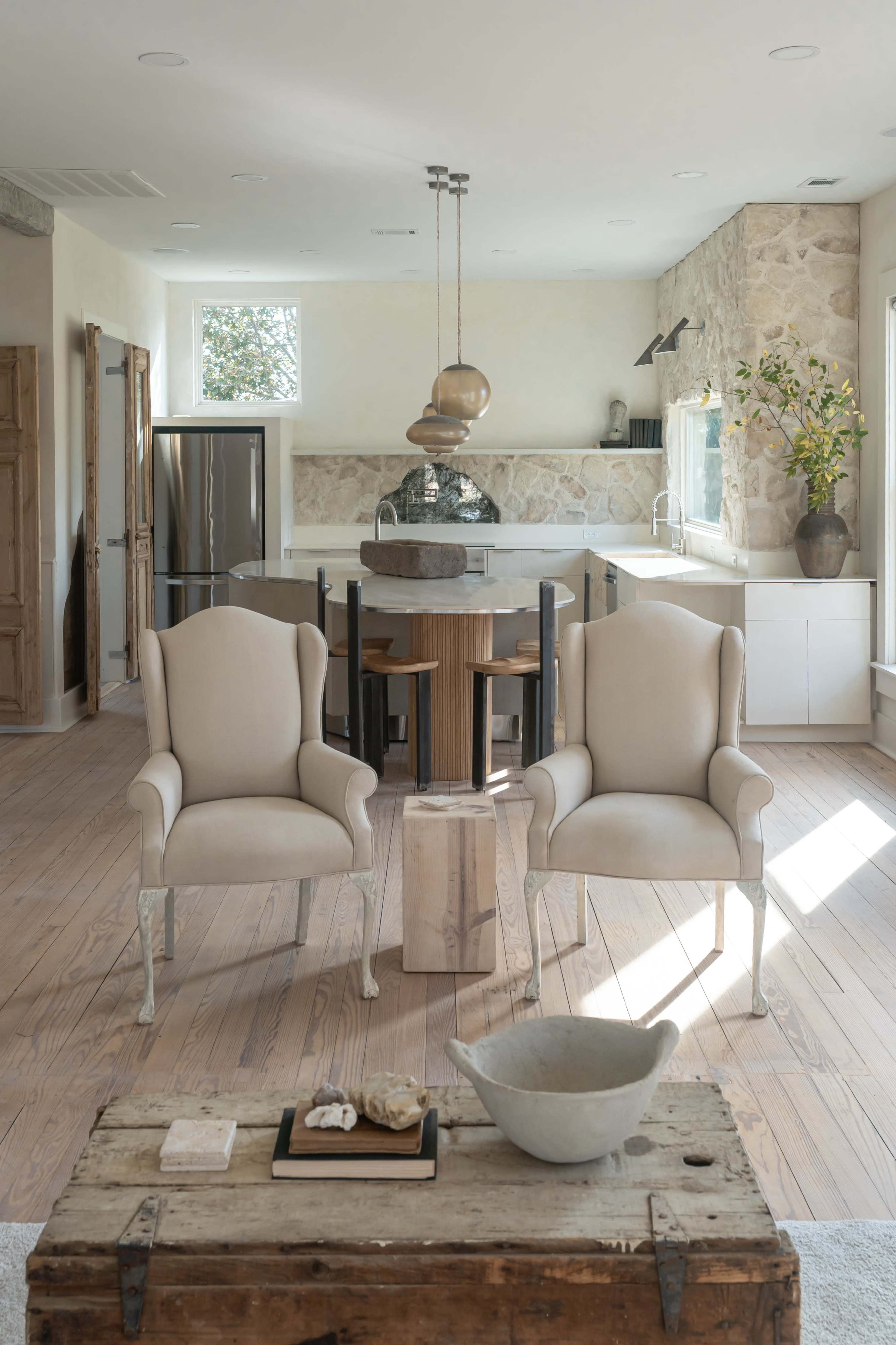 The image shows a modern kitchen and living area with two beige armchairs facing a small wooden table, stone walls, and a dining area in the background.