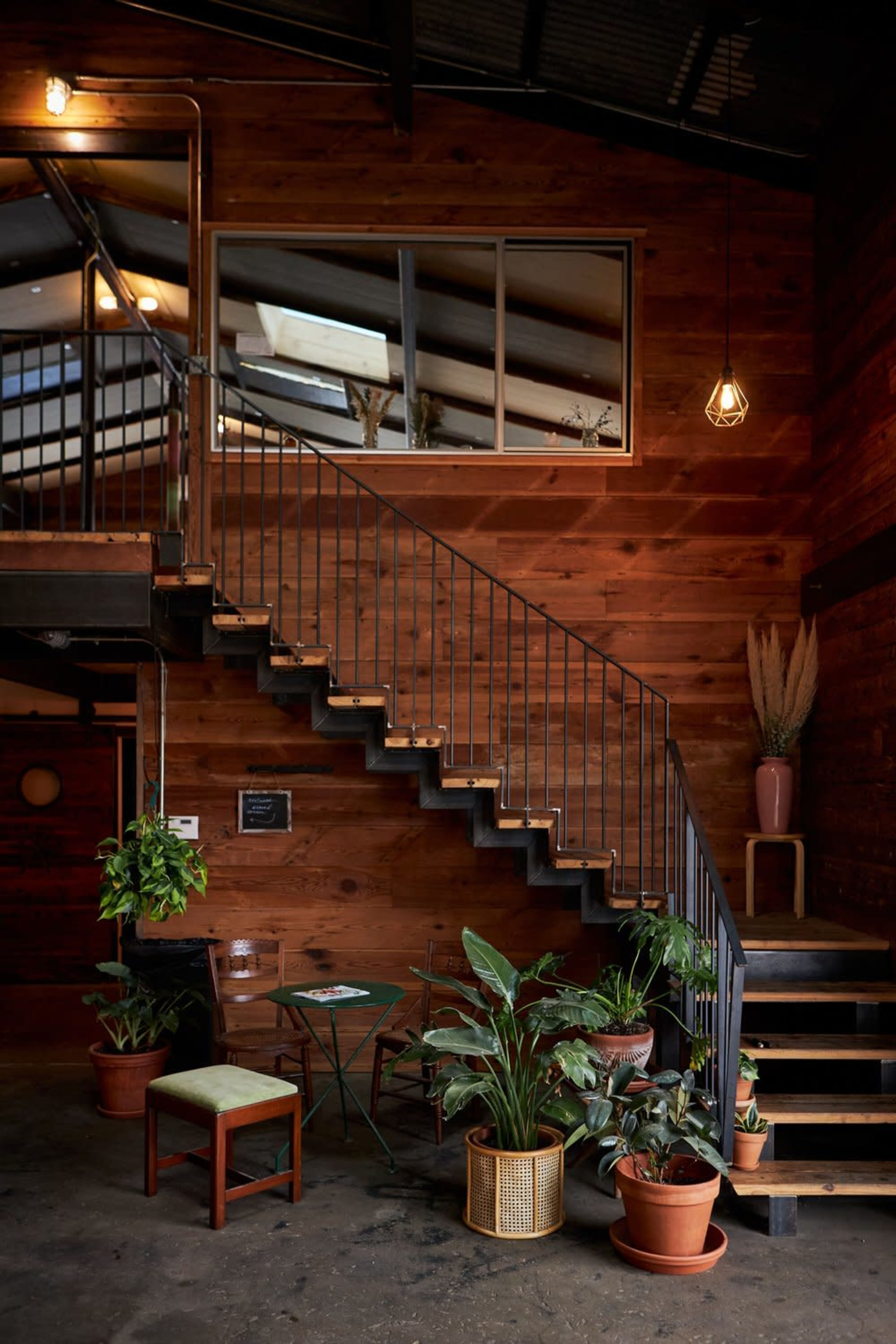 A wooden staircase with a black metal railing leads to an upper level in a room filled with potted plants and rustic decor.