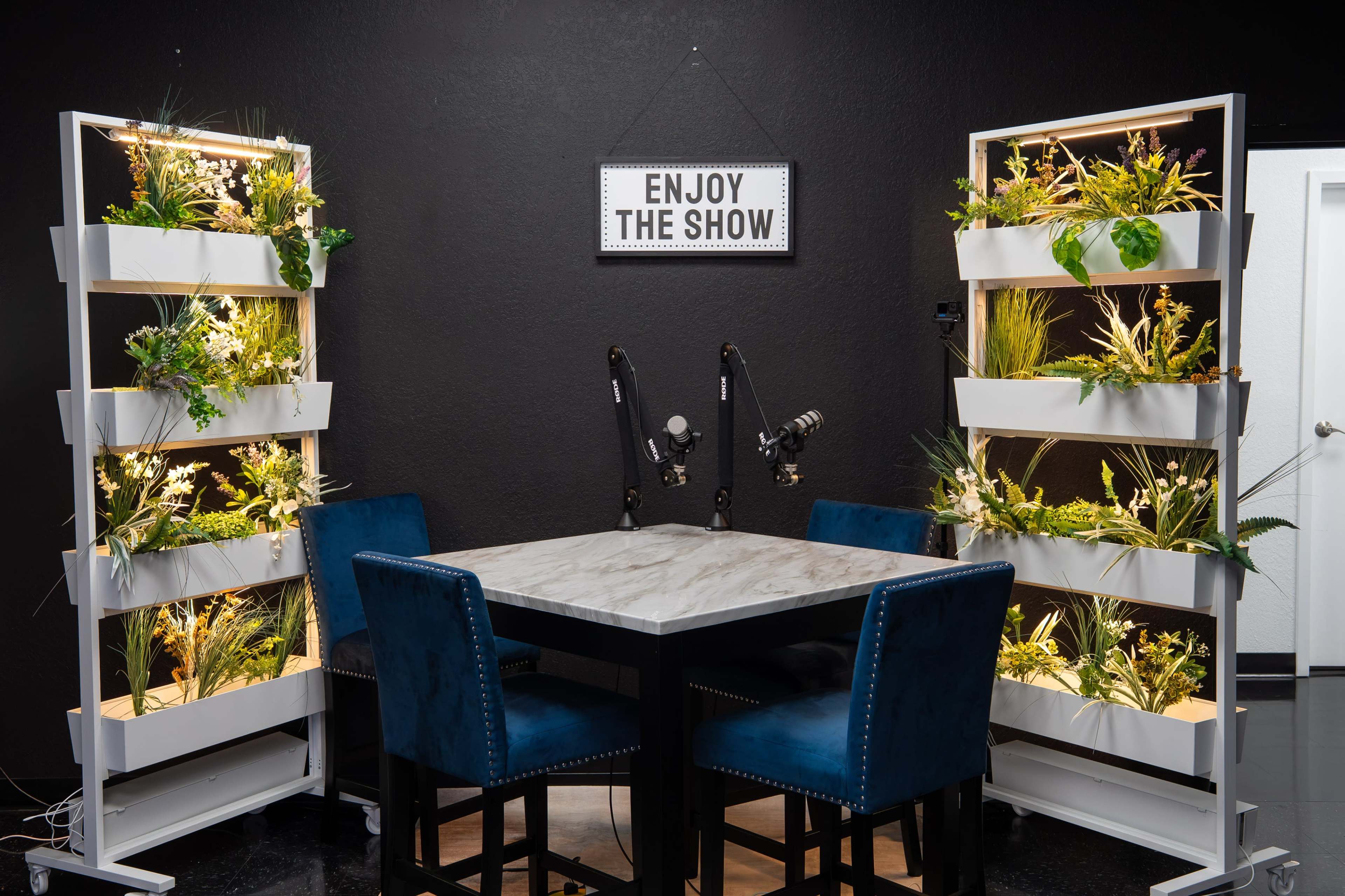 The image shows a modern workspace with a table surrounded by blue chairs, flanked by two vertical planters filled with greenery, and a sign overhead that reads "ENJOY THE SHOW."