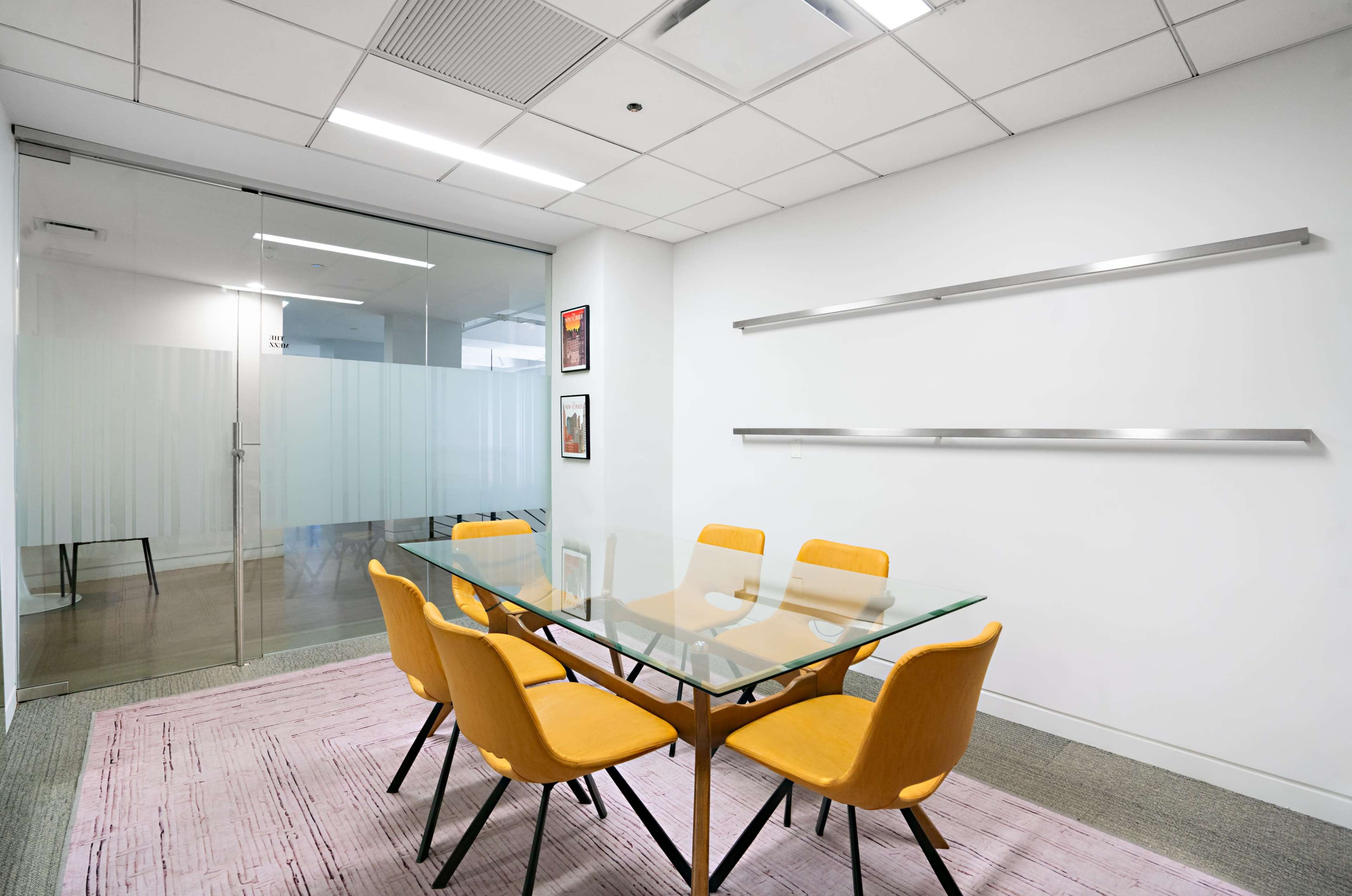 A glass conference table with four yellow chairs is positioned in a bright office meeting room featuring a glass wall and a light-colored carpet.