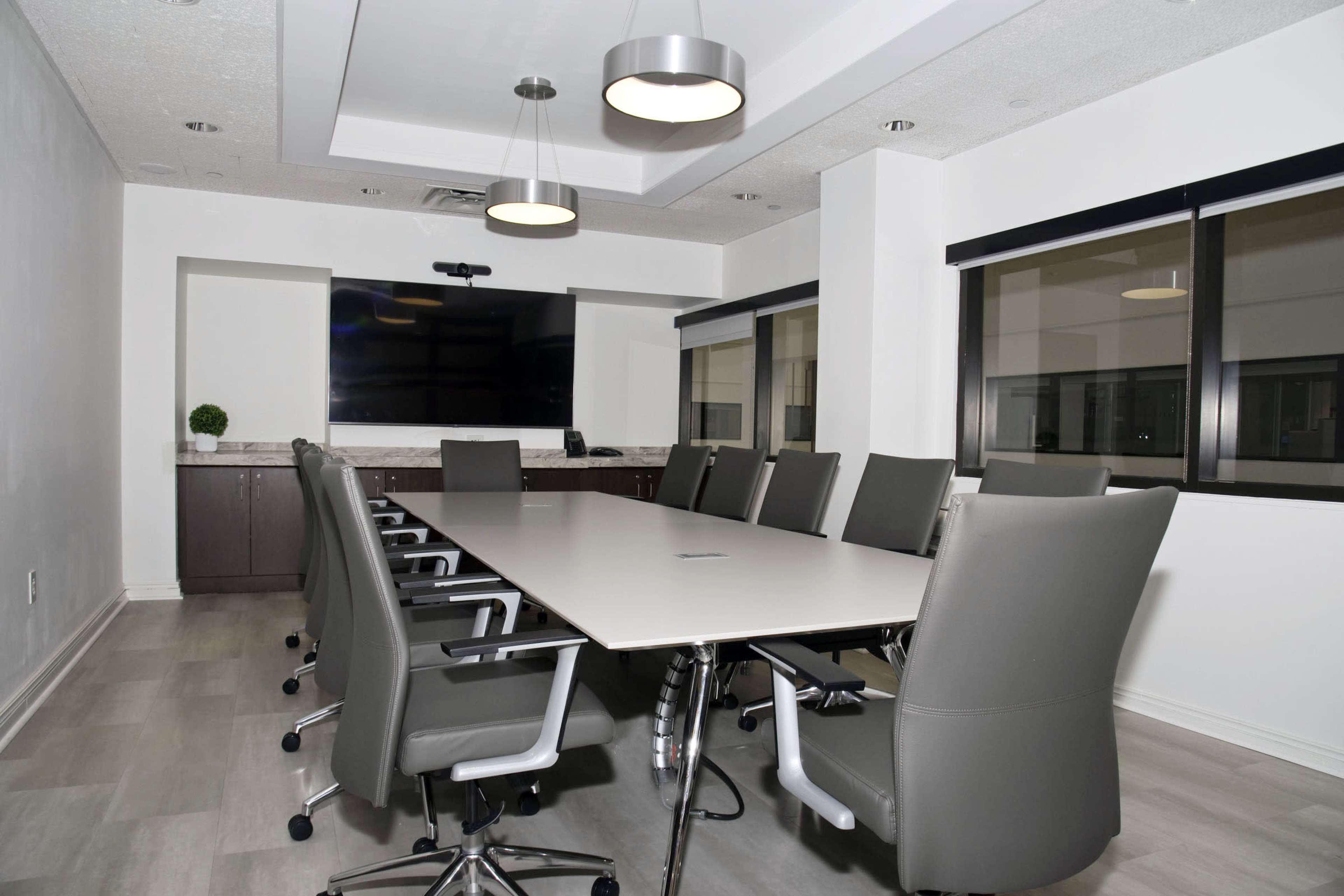The image shows a modern conference room with a long table surrounded by ergonomic chairs, a large TV screen on the wall, and windows providing natural light.