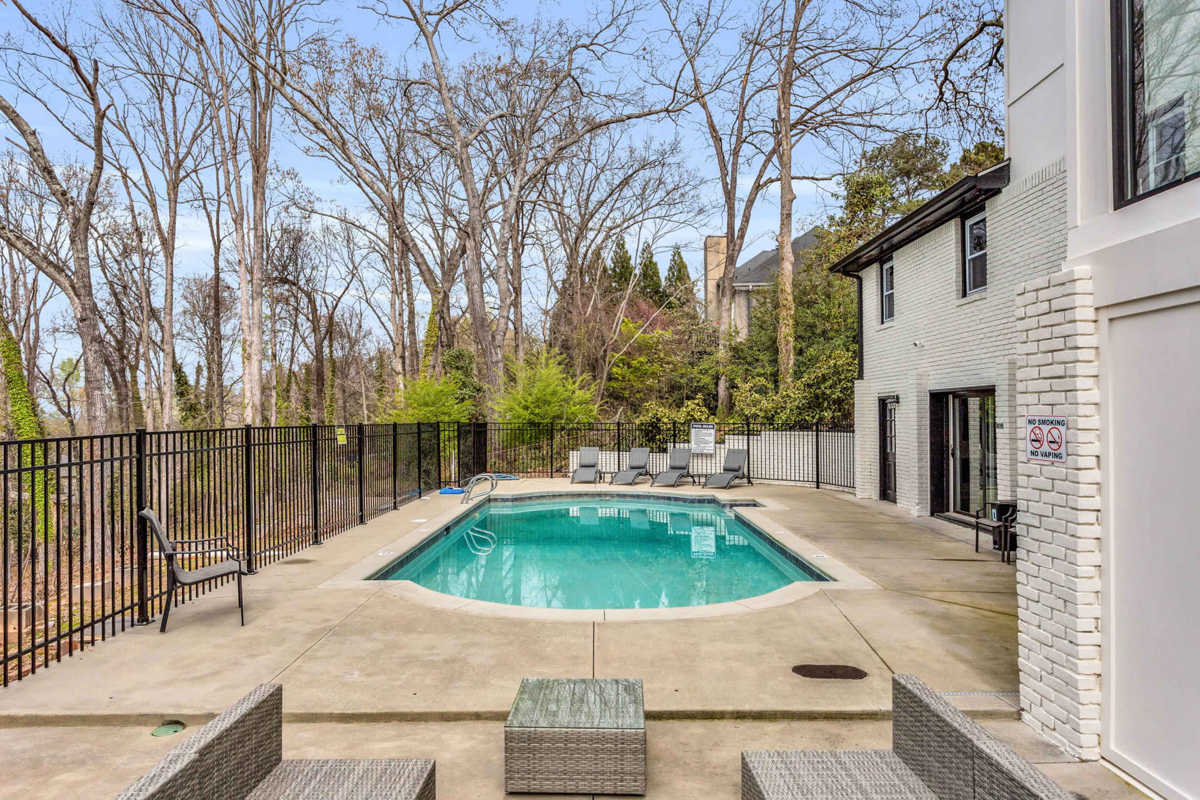 A backyard pool area surrounded by tall trees, with lounge chairs and a fence enclosing the pool.