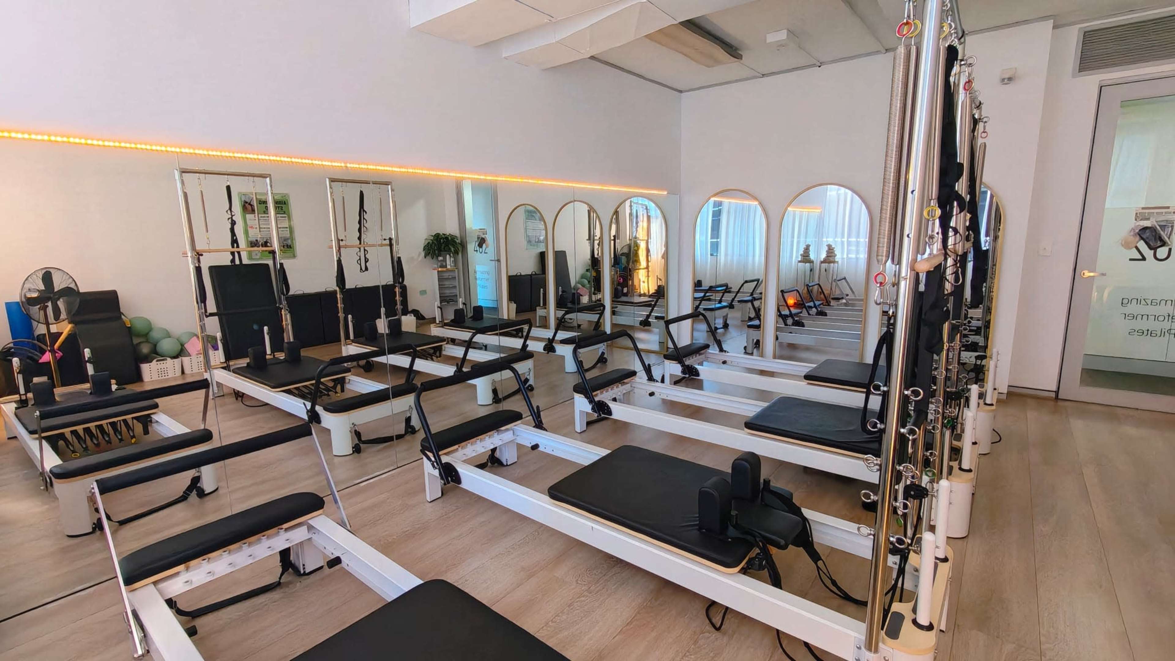 The image shows a bright and spacious Pilates studio featuring several reformer machines arranged neatly in a clean, modern interior.