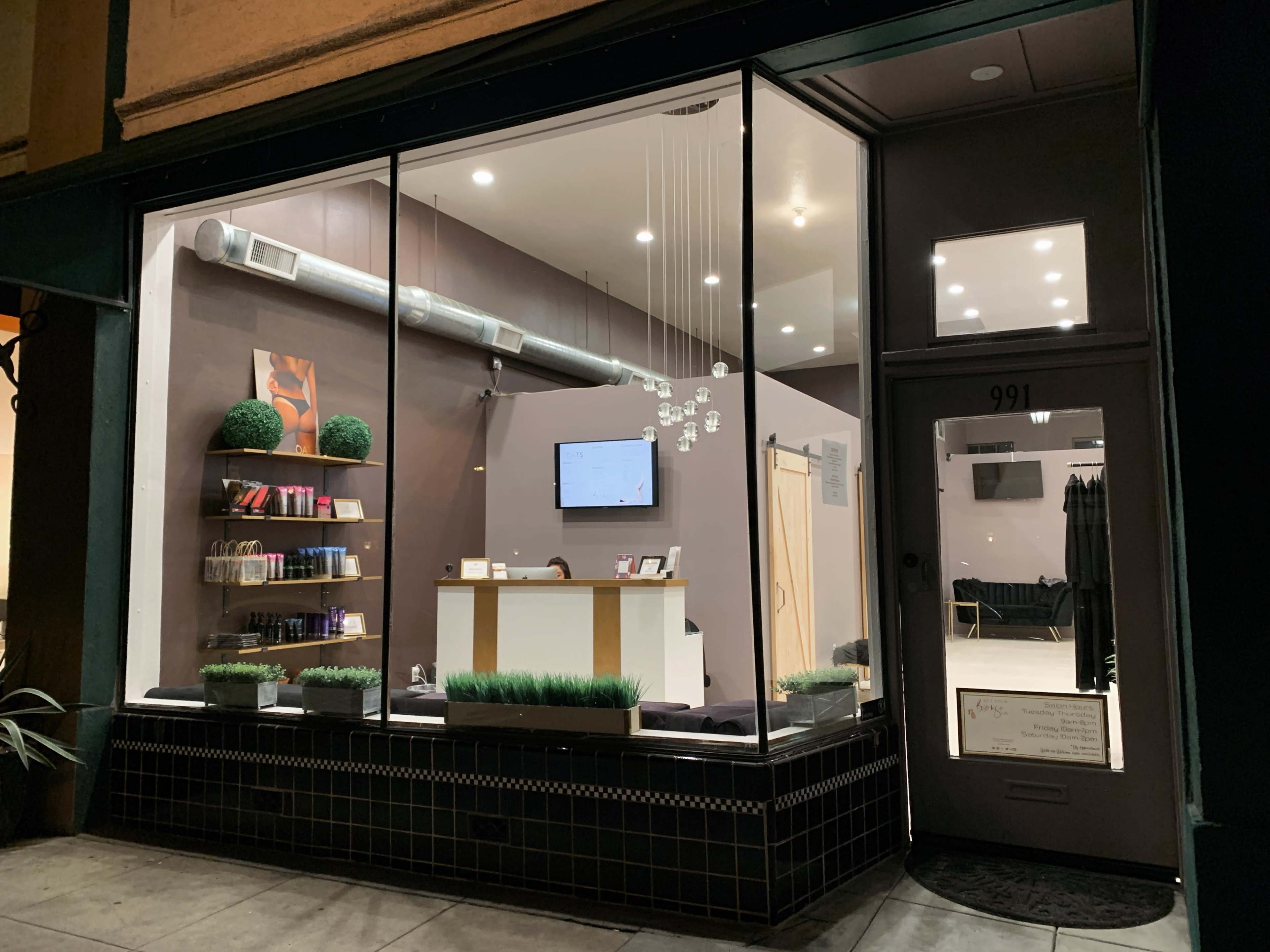 The image shows a modern storefront featuring large glass windows, a minimalistic interior with a reception desk, shelves displaying products, and decorative plants.