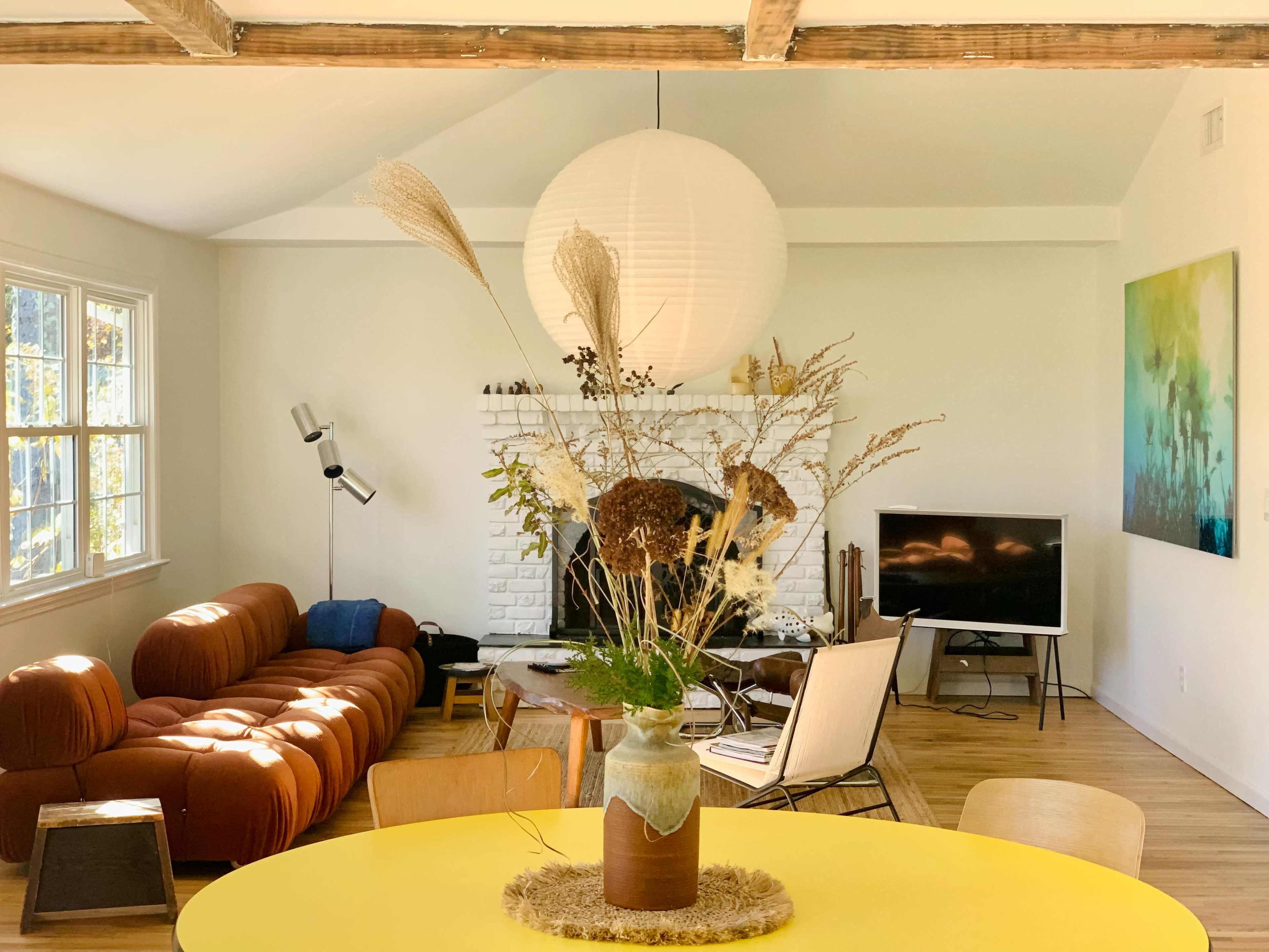 A spacious living room features a round yellow table with a vase of dried plants, a brick fireplace, a modern sofa, and a television.