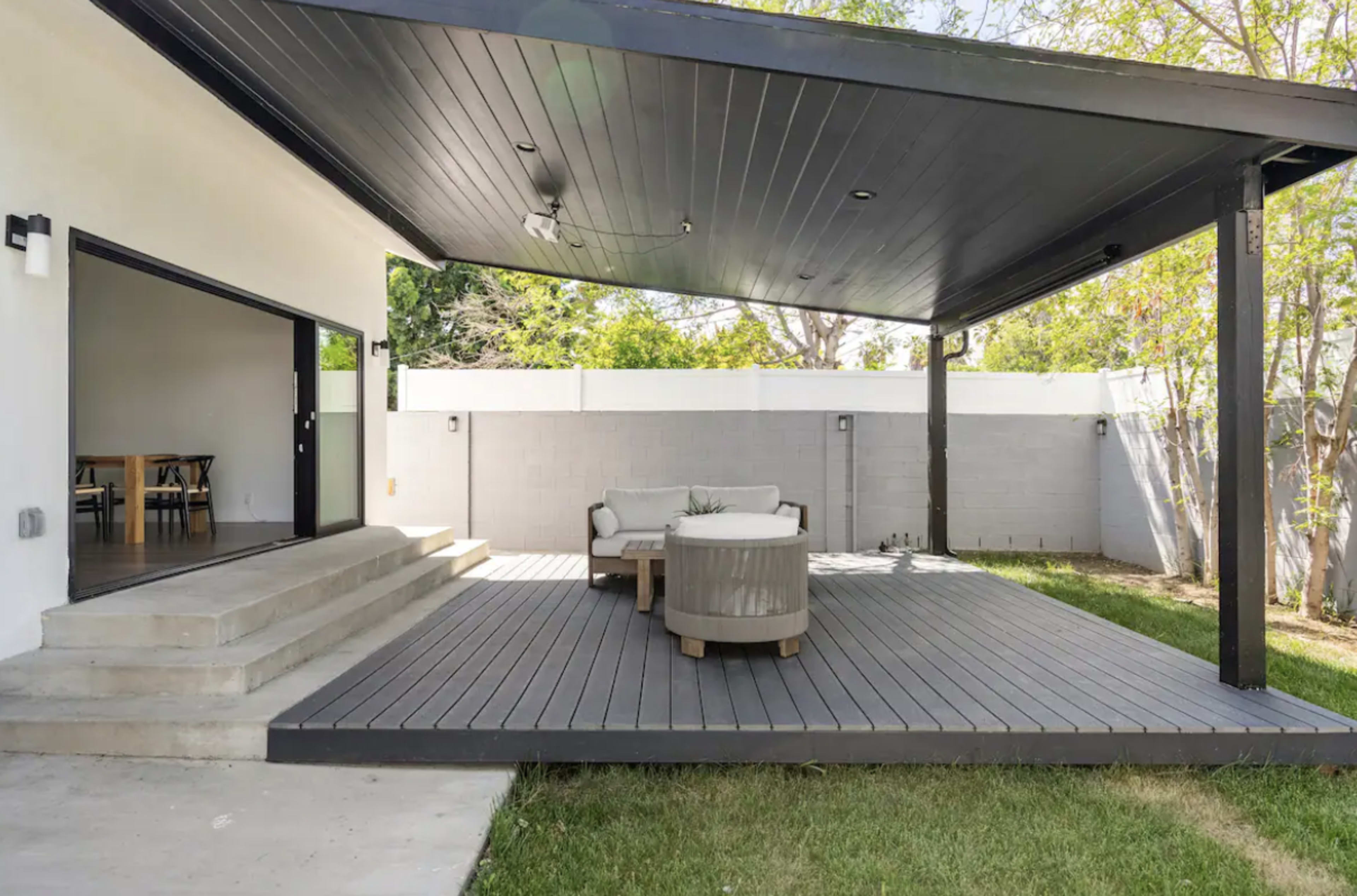 A modern outdoor patio with a shaded seating area, wooden deck, and a grassy yard bordered by a wall.
