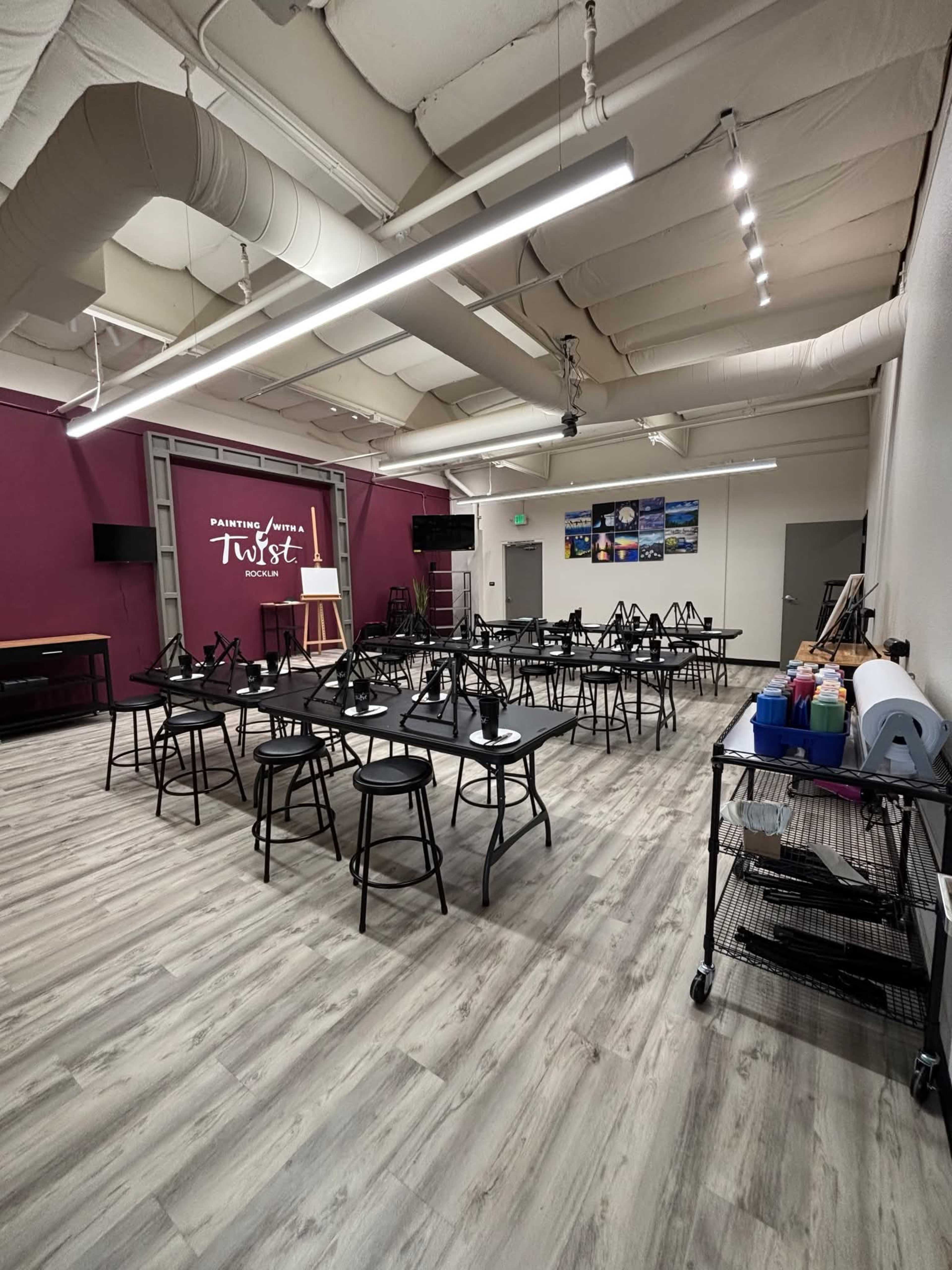 The image shows a spacious art studio with several black tables and stools arranged for a class, along with easels and supplies ready for use.