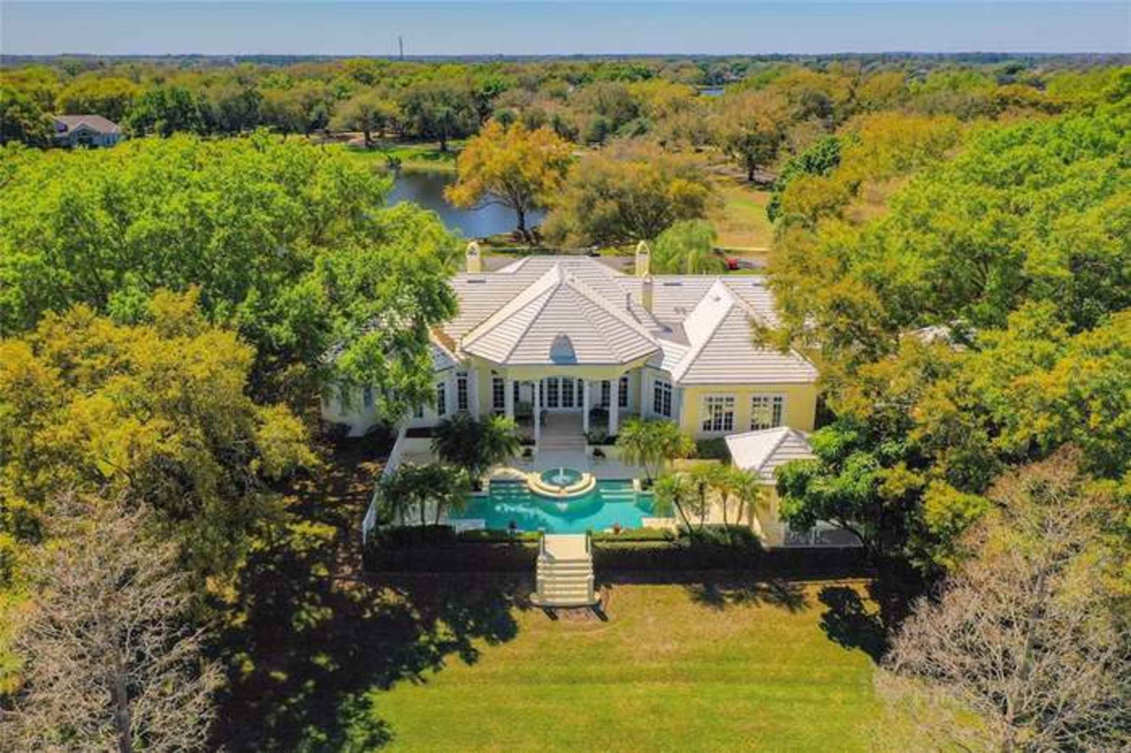 A spacious luxury home with a pool surrounded by trees and a pond in the background.