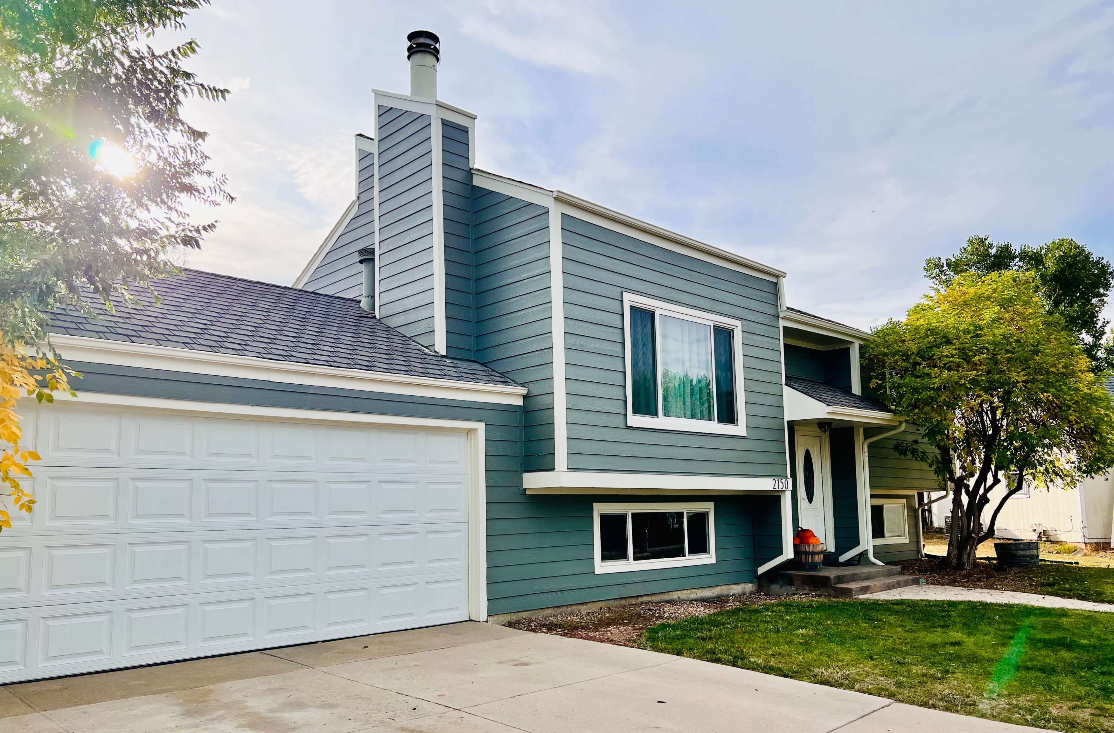 A two-story house with a blue-green exterior, a prominent chimney, and a spacious garage is set on a well-maintained lawn.