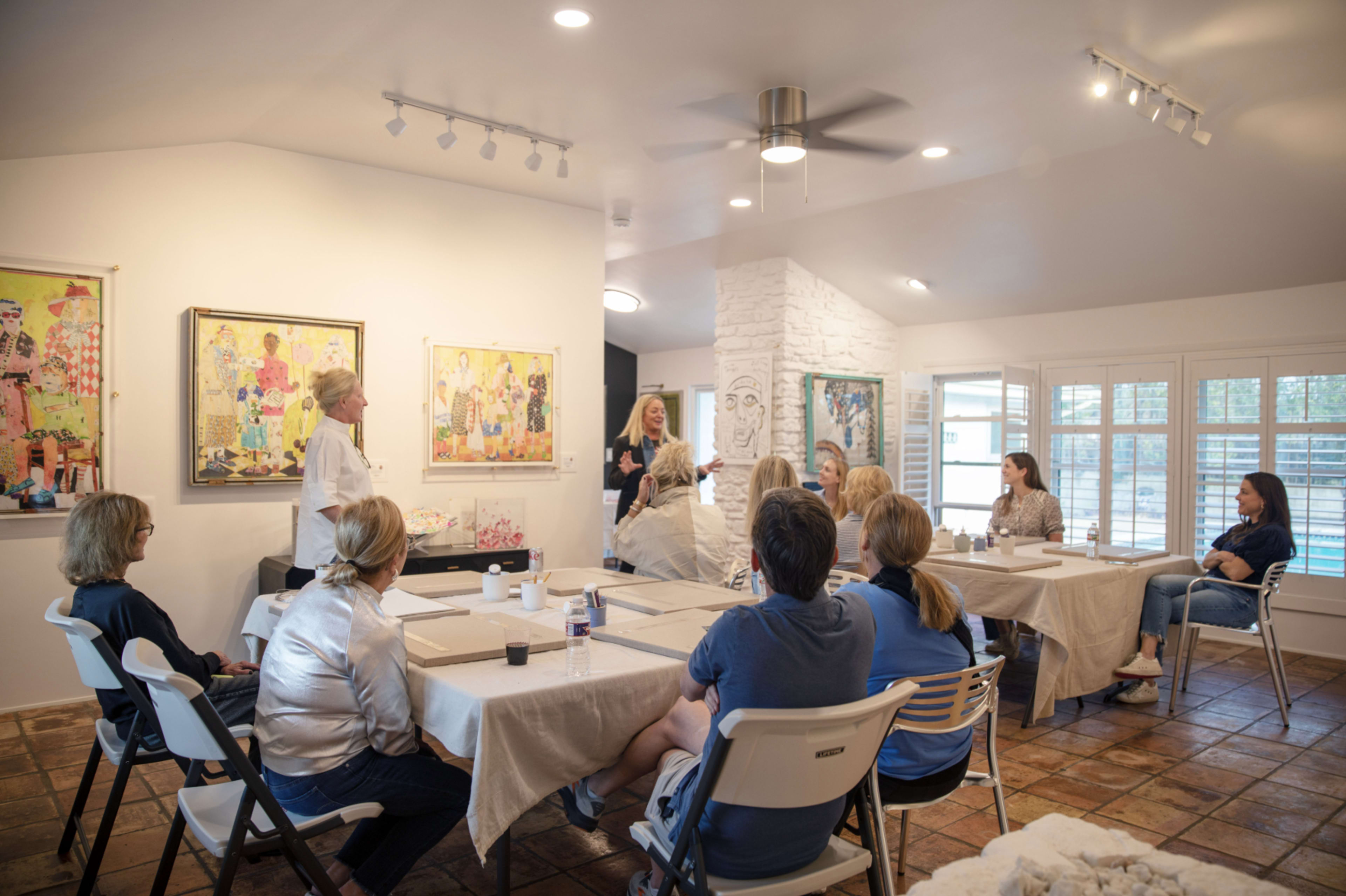 A group of people is seated at tables in a well-lit art studio, attentively listening to a speaker who stands at the front near colorful artwork on the walls.