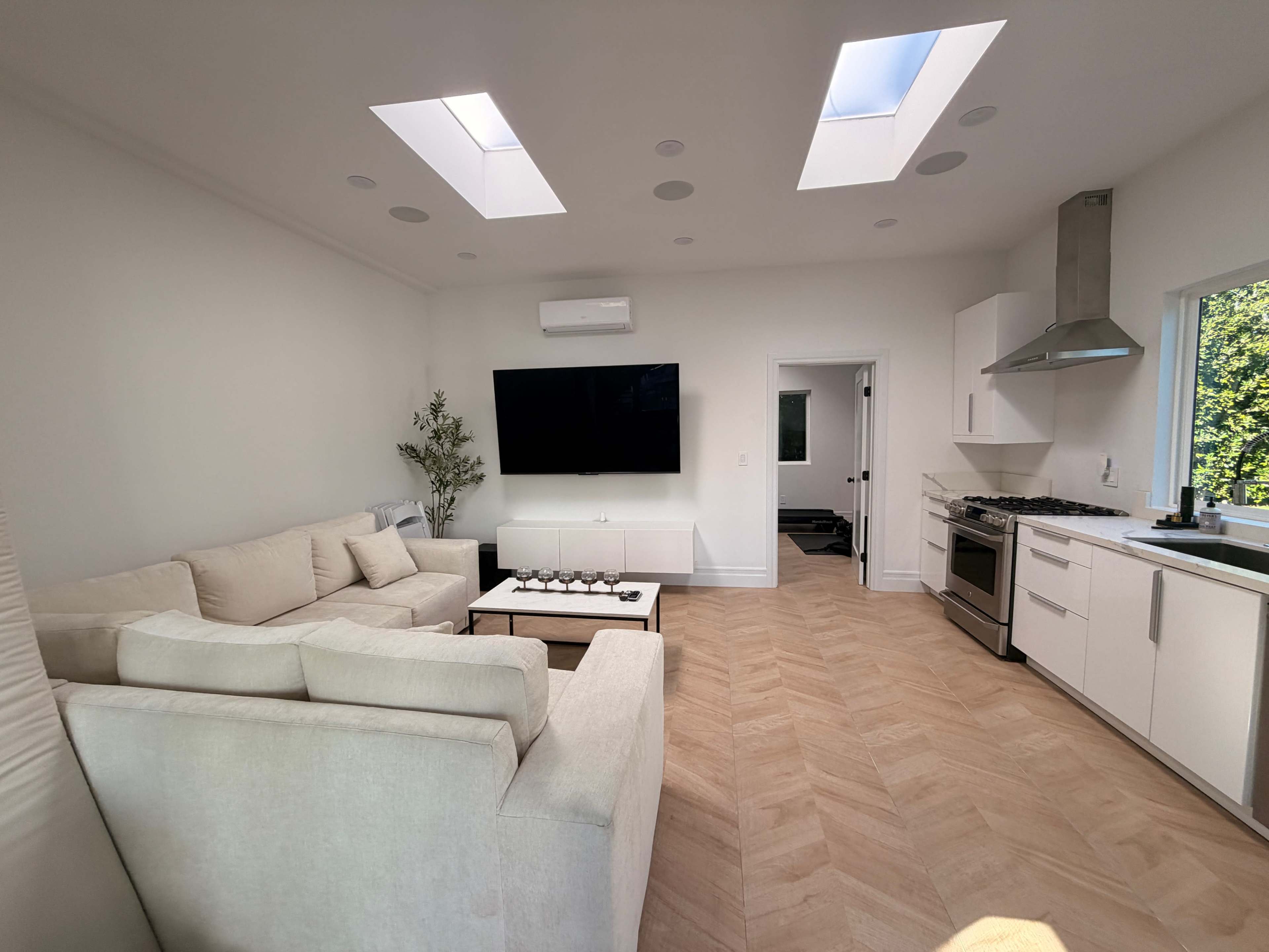 The image shows a modern living room with a large sectional sofa, a flat-screen TV mounted on the wall, and a well-equipped kitchen area with skylights and a dining table.