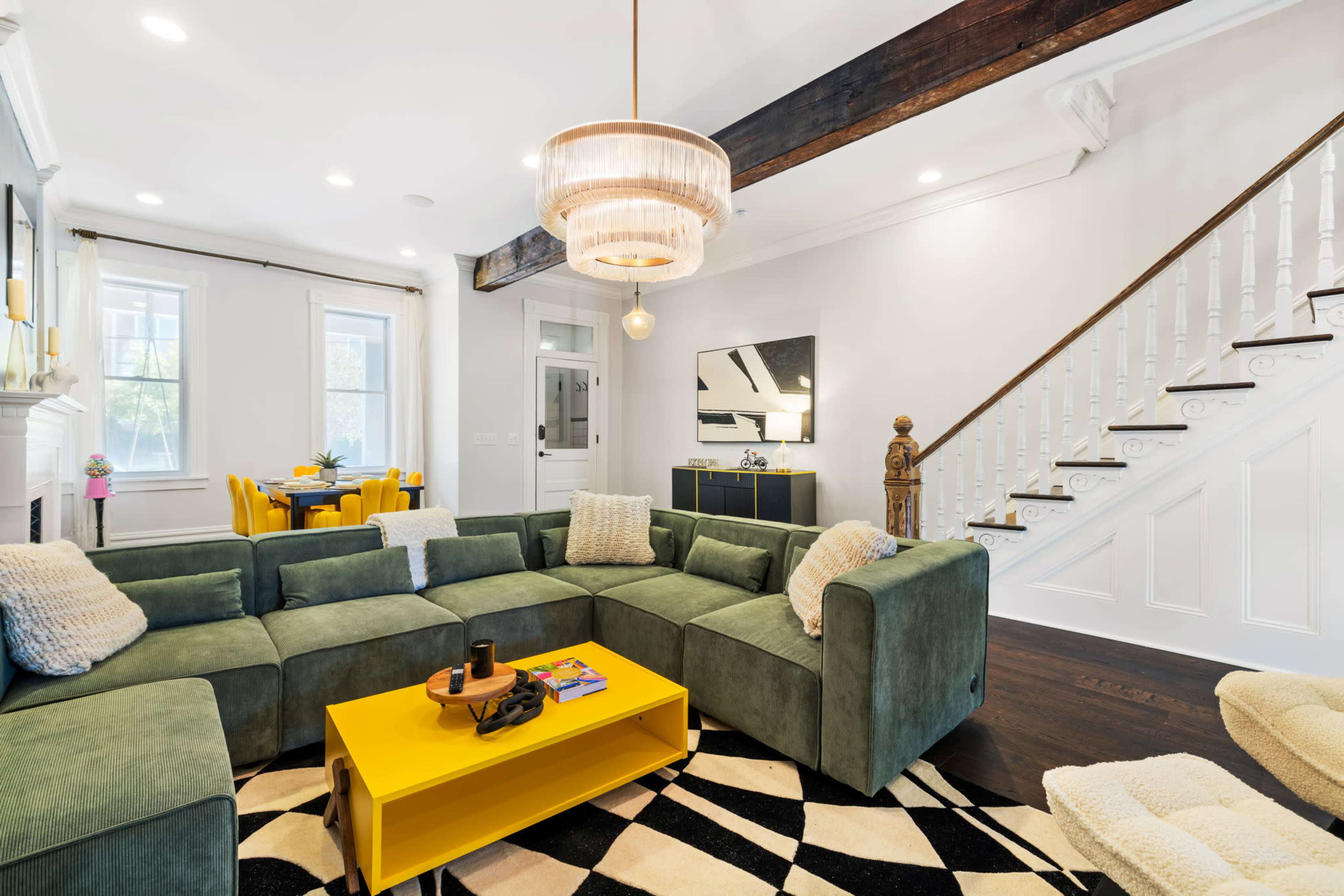 The living room features a large, green sectional sofa arranged around a yellow coffee table, with a staircase and a dining area visible in the background.
