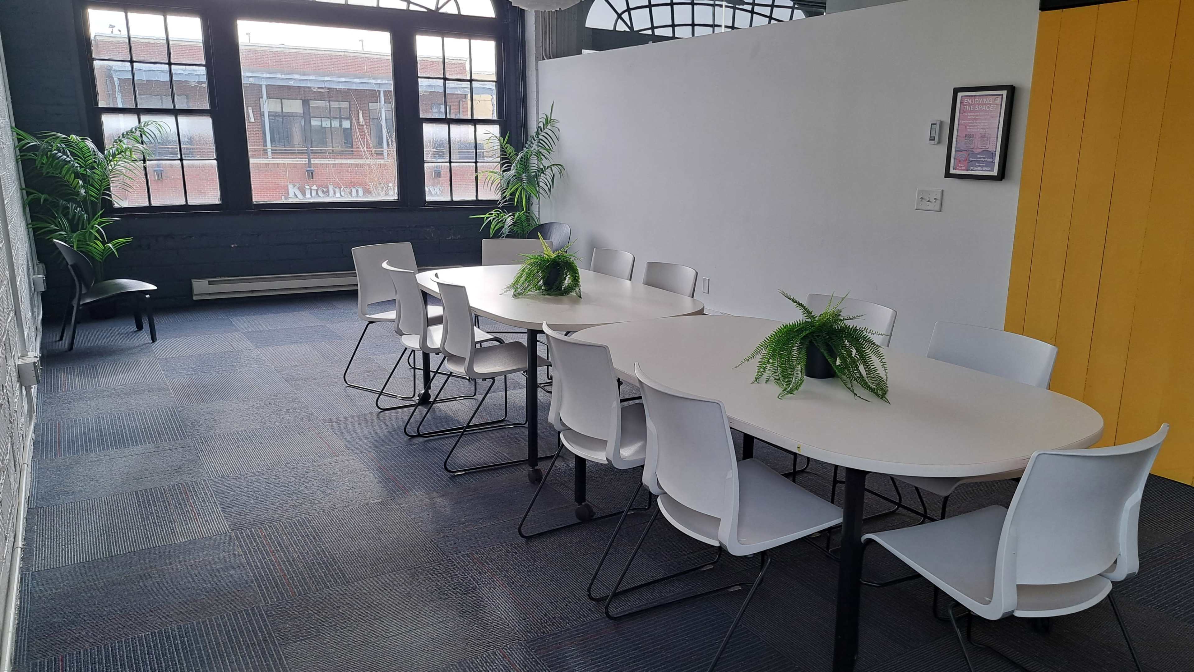 A long, white table with several white chairs is arranged in a well-lit room featuring large windows and decorative green plants.