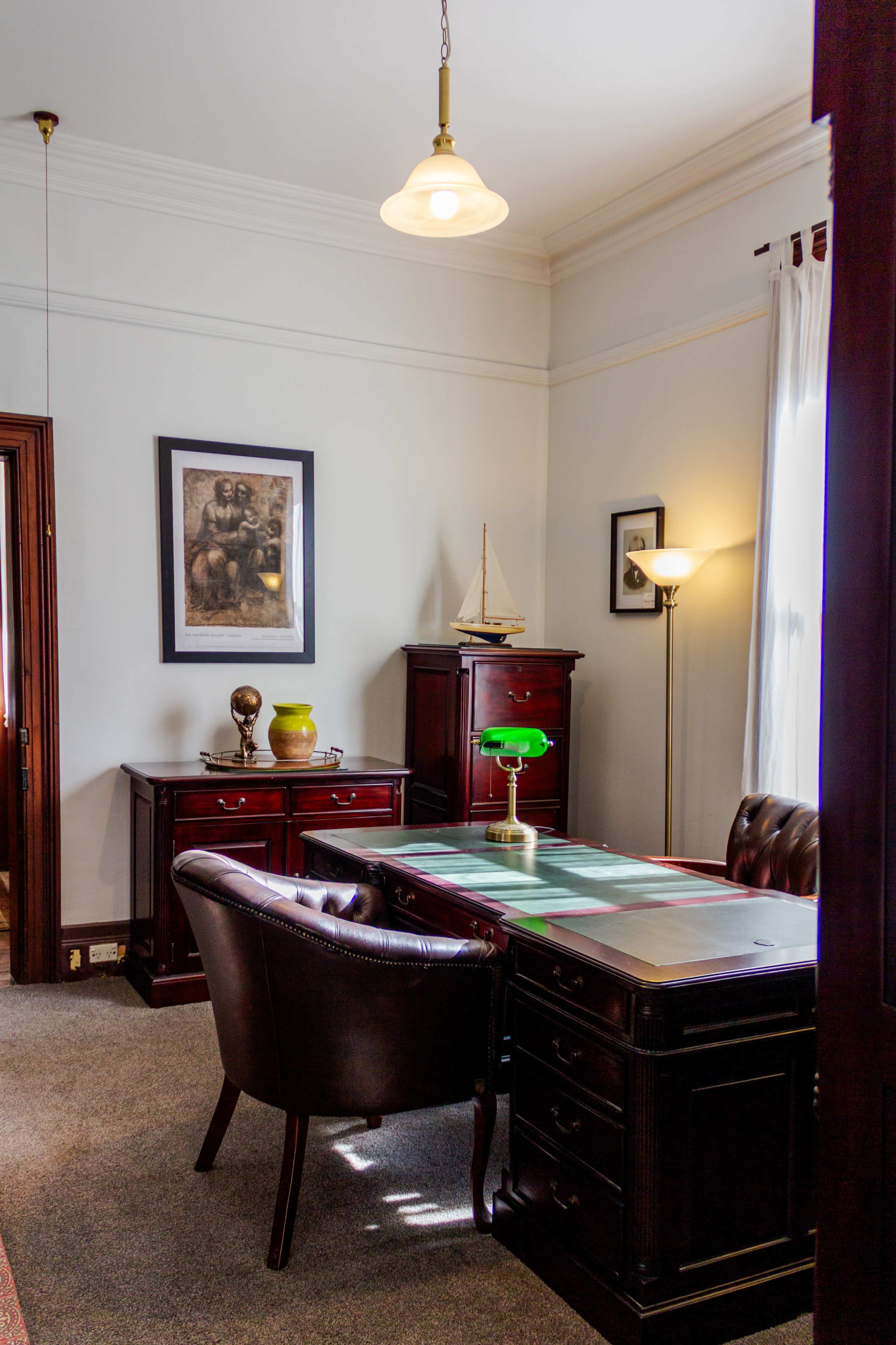The image shows a classic office space featuring a wooden desk, a leather chair, a green desk lamp, and dark wood furniture, with a framed artwork on the wall.