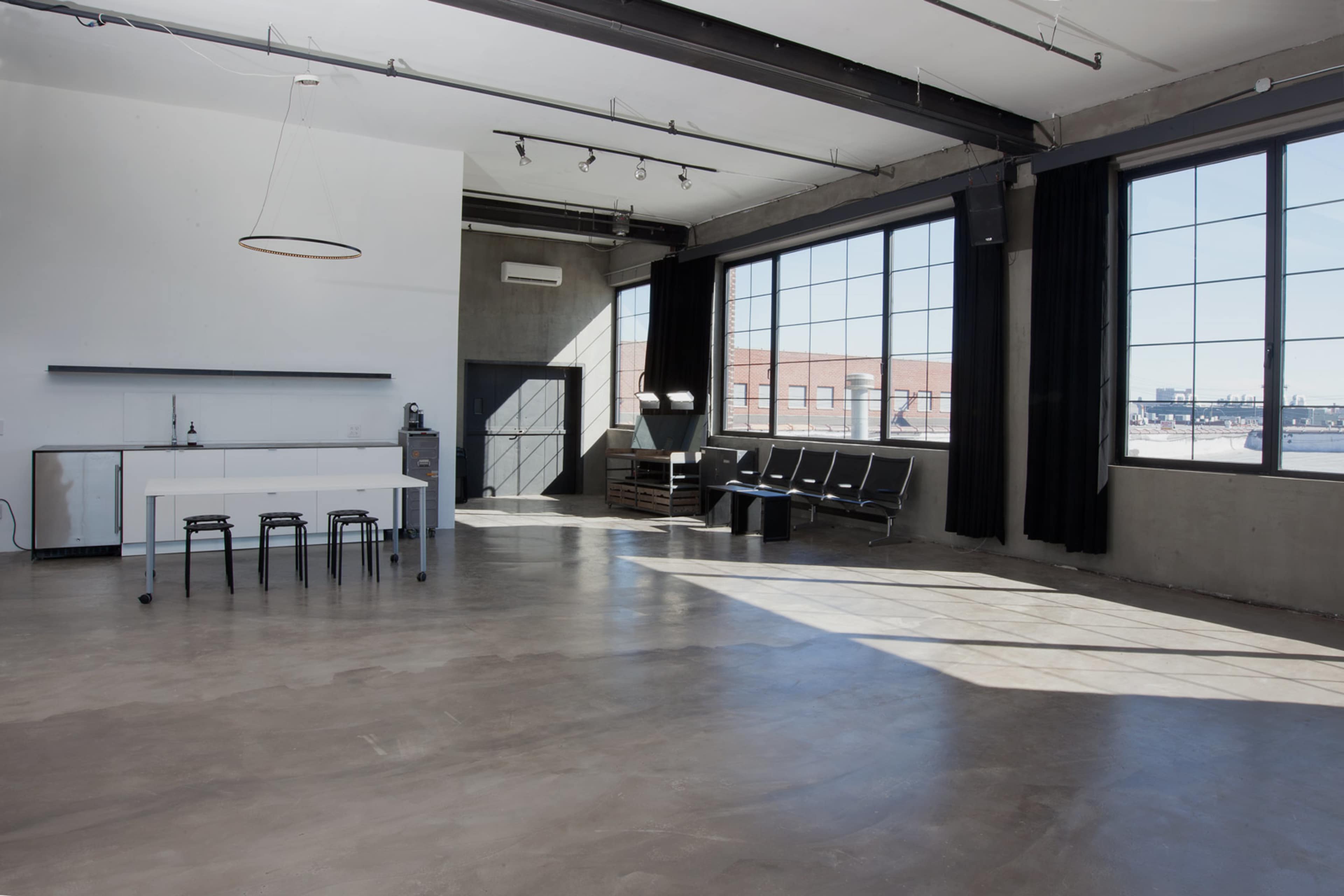 The image shows a modern, open space with large windows, minimal furniture, and a kitchenette area.