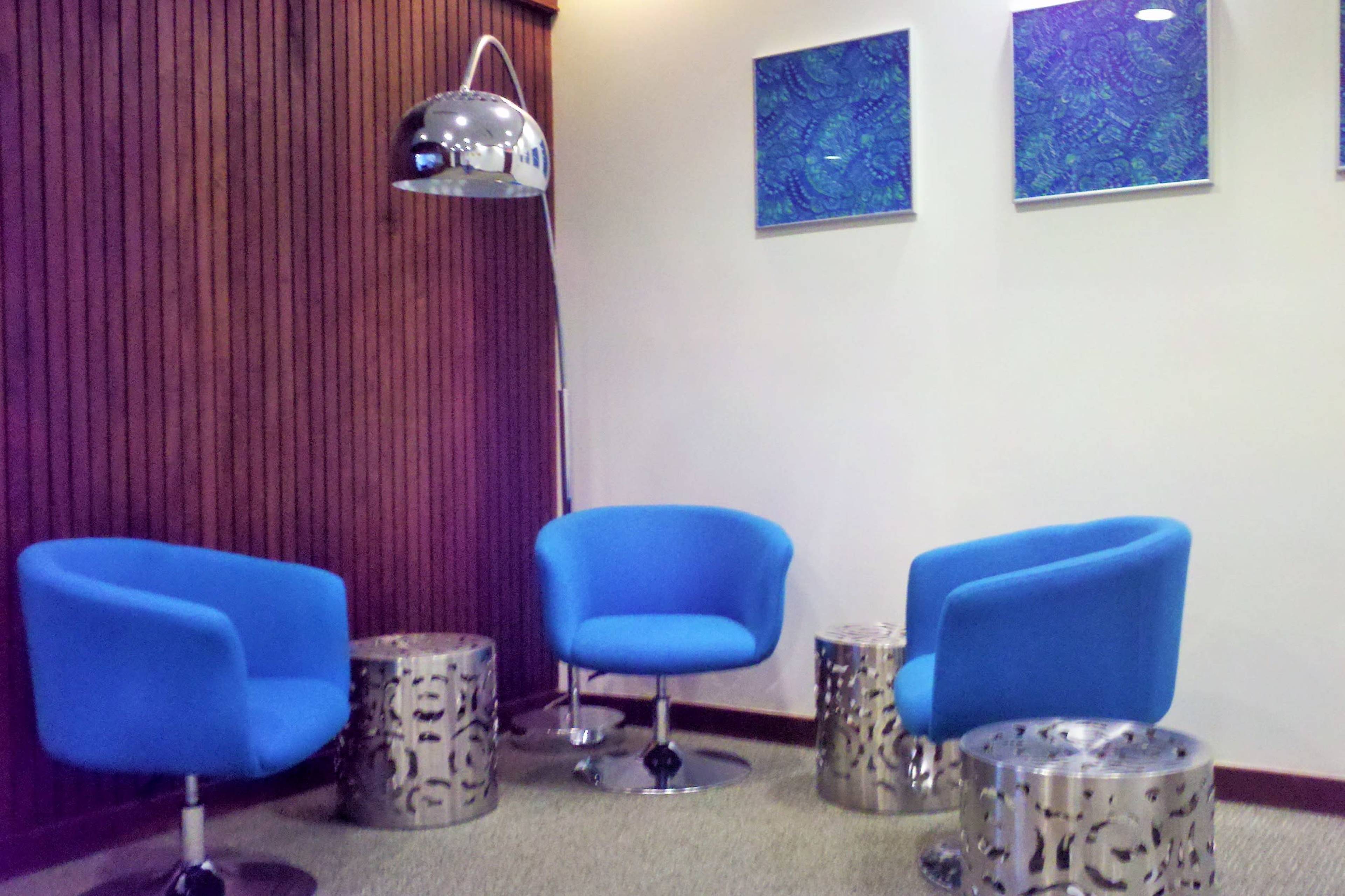 The image shows a small lounge area with three blue chairs and two metallic side tables, accompanied by a tall lamp and colorful artwork on the wall.