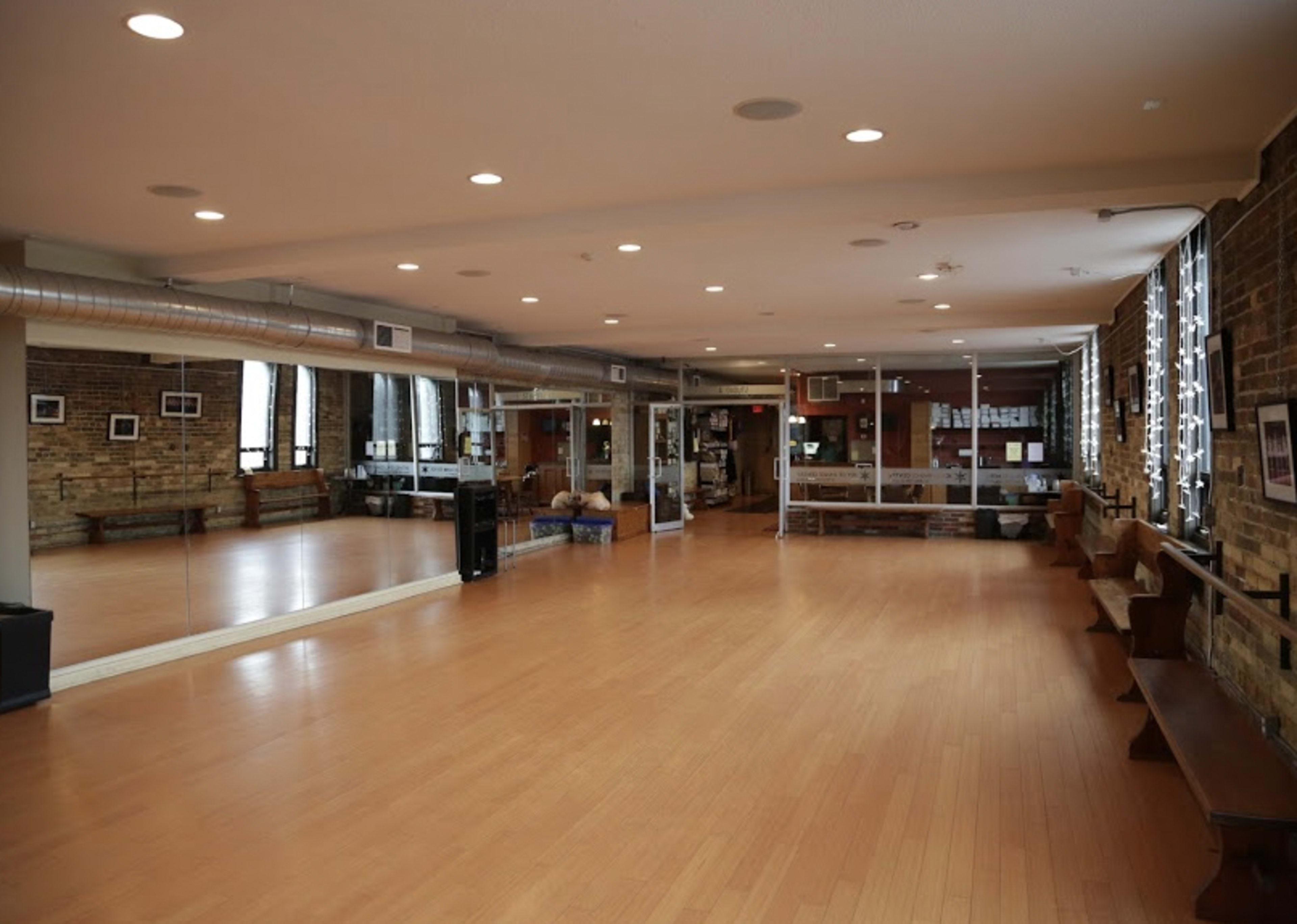 The image shows a spacious dance studio with wooden floors, large mirrors on one wall, and exposed brick walls, illuminated by overhead lighting.