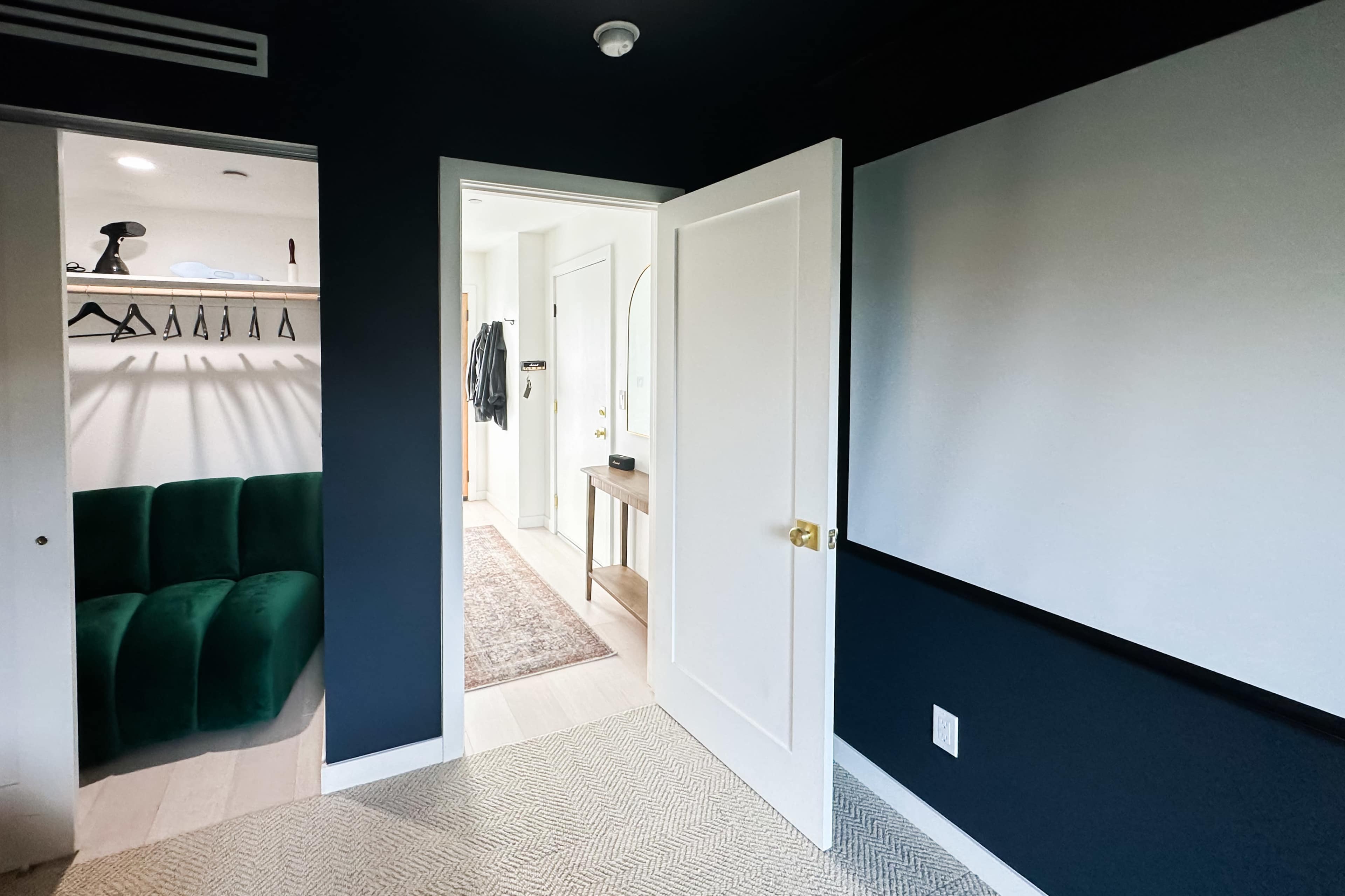 A doorway opens into a dark-walled room featuring a green sofa, a screen on the wall, and a closet with hanging space.