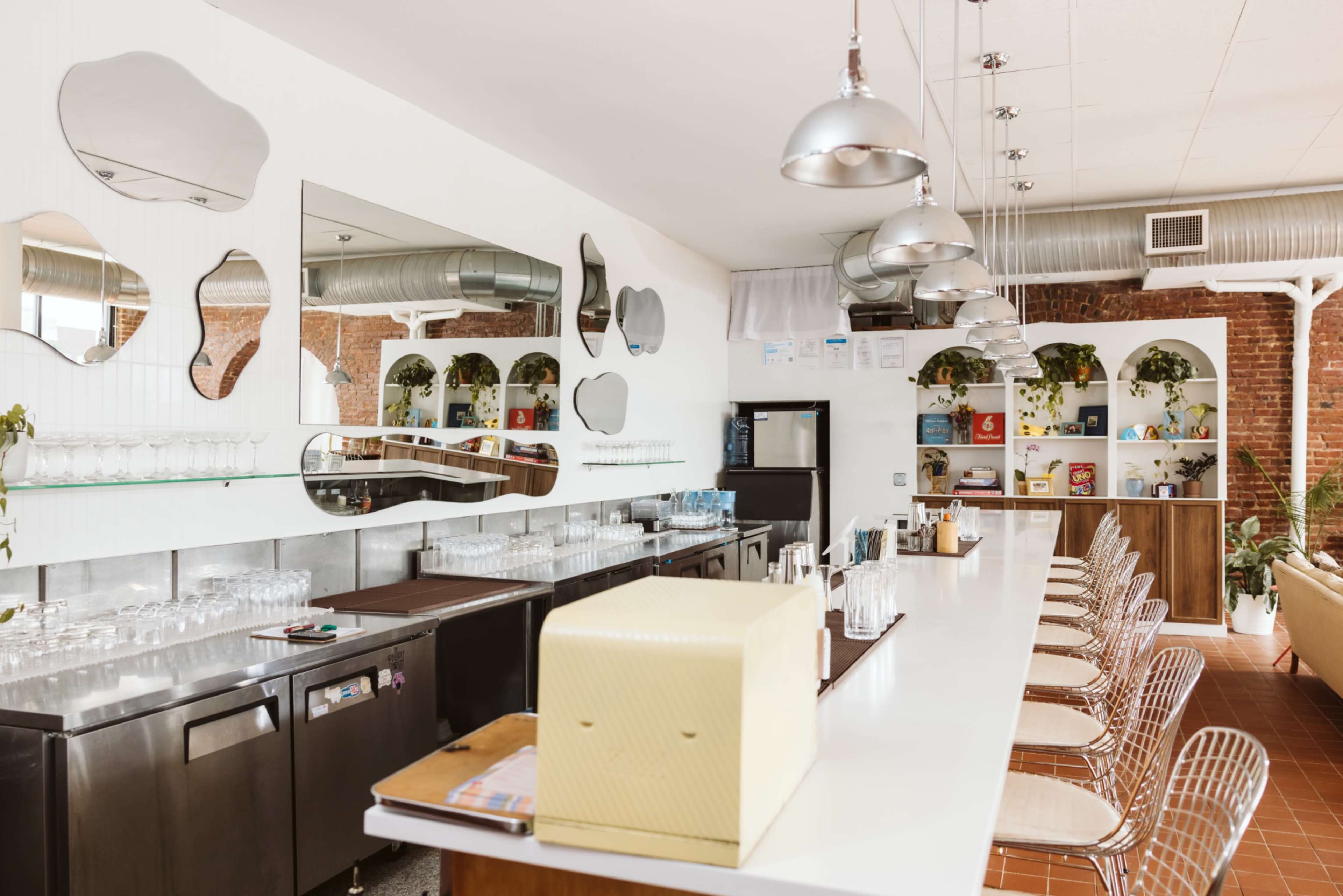The image shows a modern café bar with a white countertop, metal pendant lights, and shelves displaying various items against a brick wall backdrop.