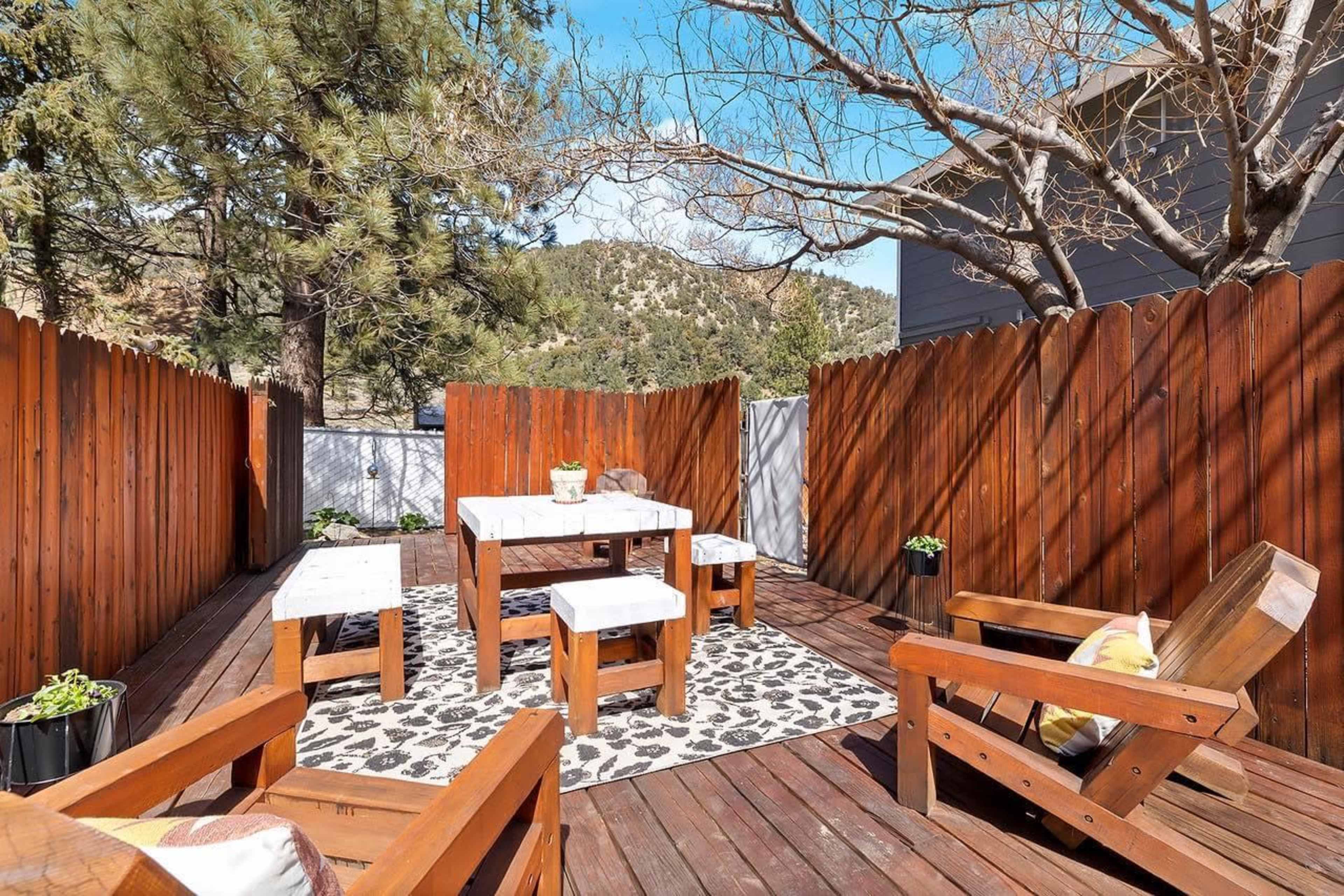 A wooden deck features a table surrounded by four chairs, with a patterned rug beneath and a fenced background of trees and hills.