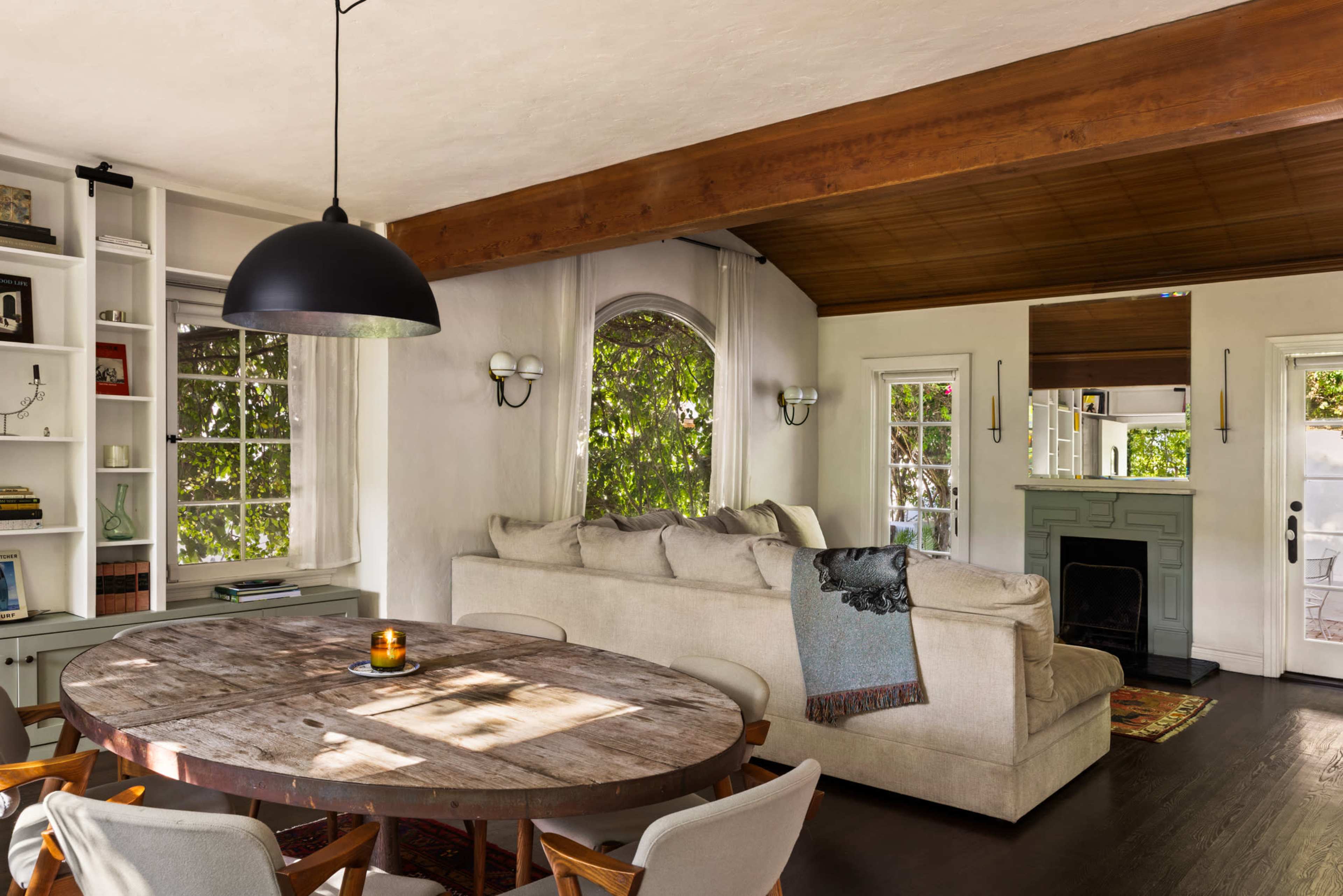 A comfortable living area features a wooden dining table, a plush sofa, and large windows allowing natural light to enter.