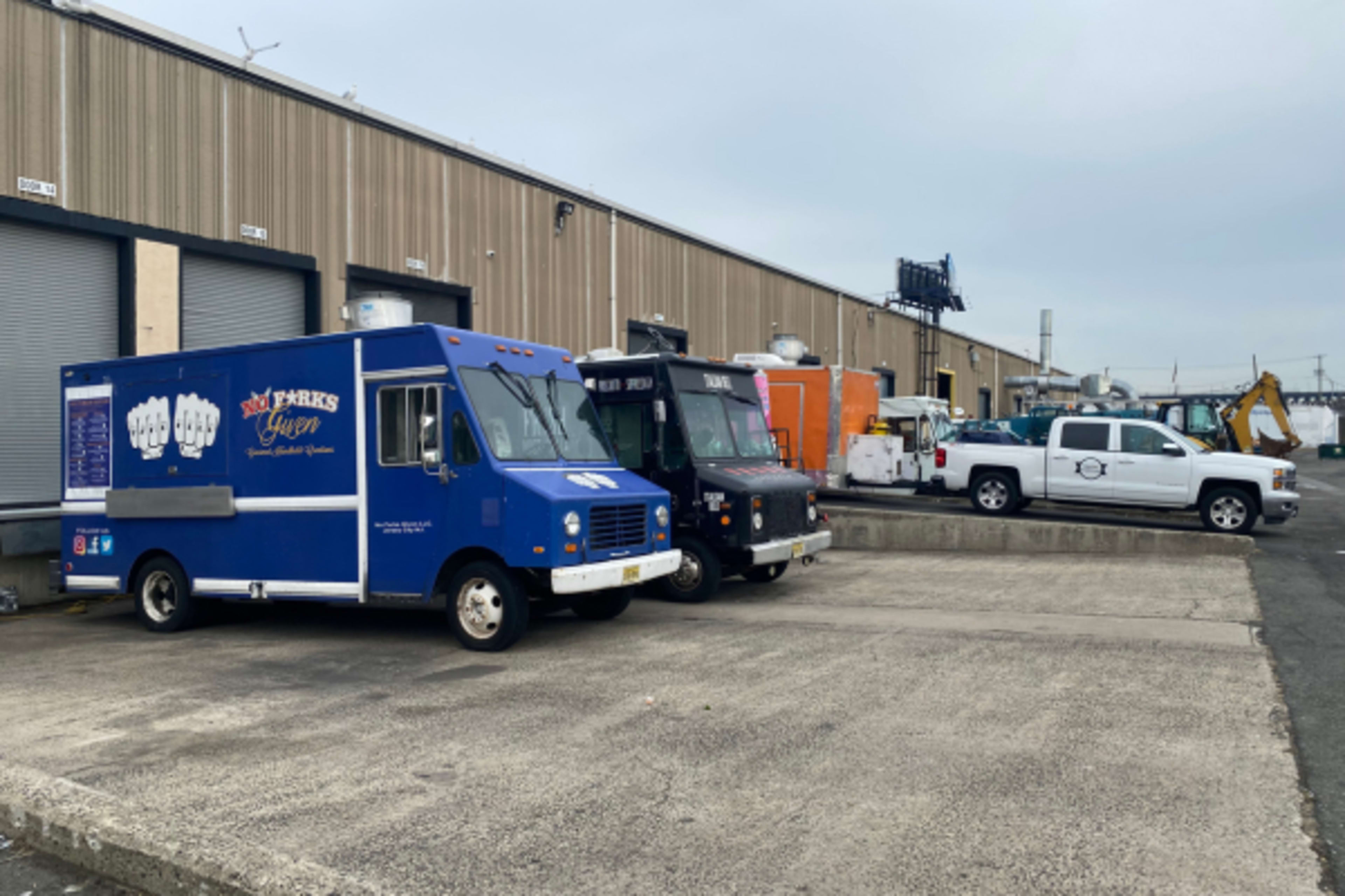 Several food trucks are parked alongside a loading dock, with a pickup truck and various other vehicles in the background.