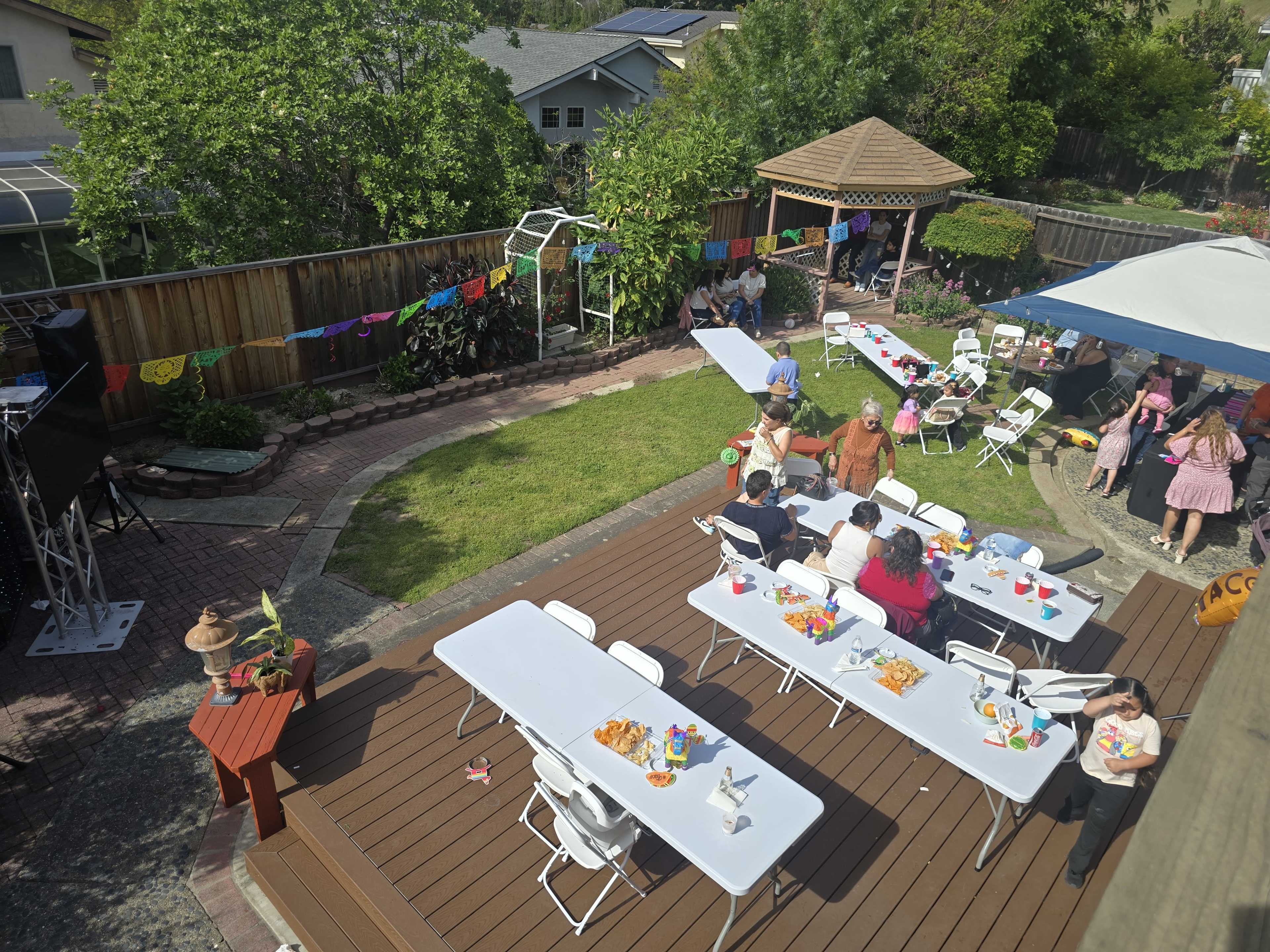 A backyard party is set up with tables and chairs, colorful decorations, and people mingling in a well-maintained garden.