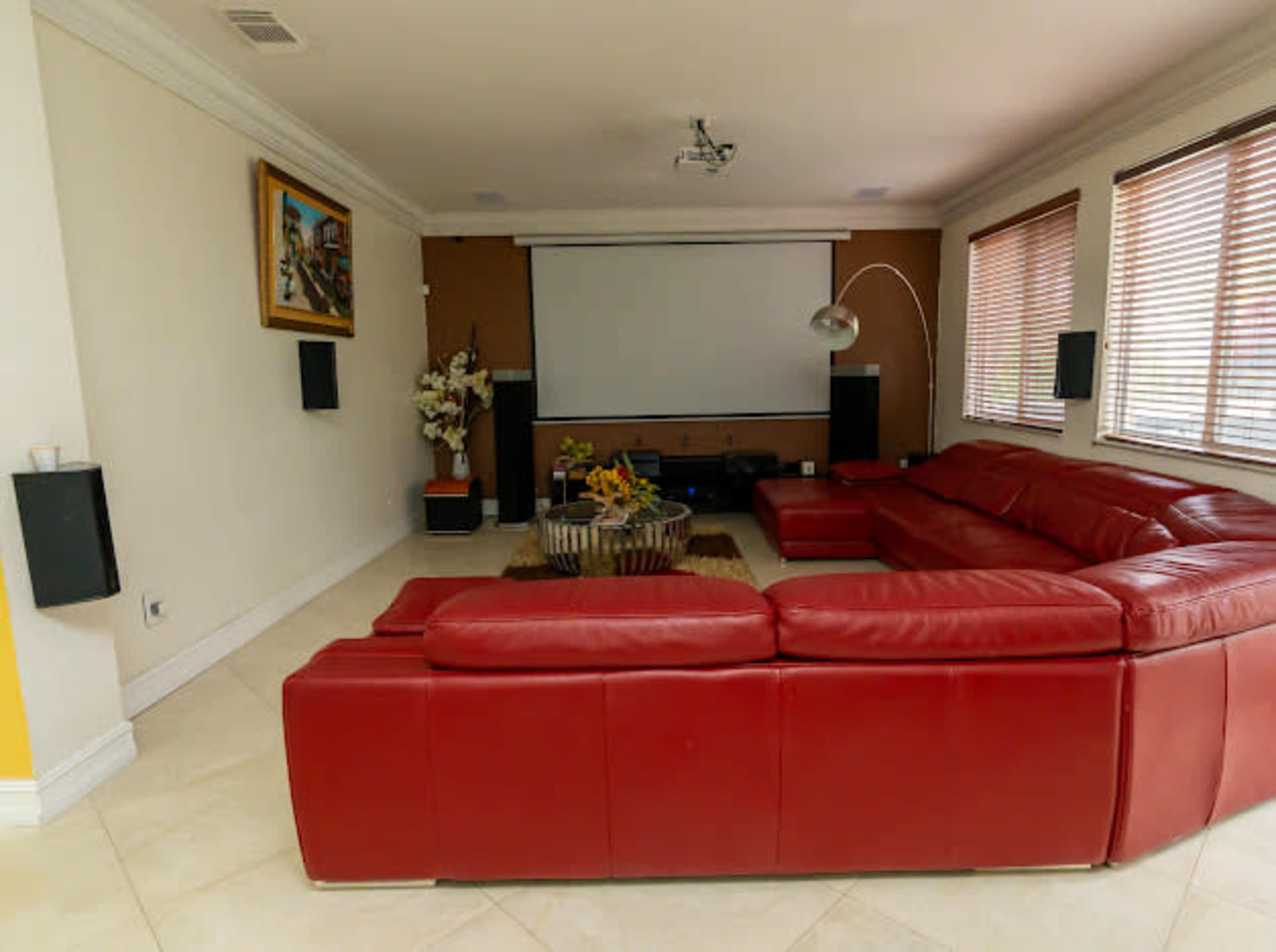 A modern living room features a large red sectional sofa, a wall-mounted projector screen, and natural light filtering through window blinds.