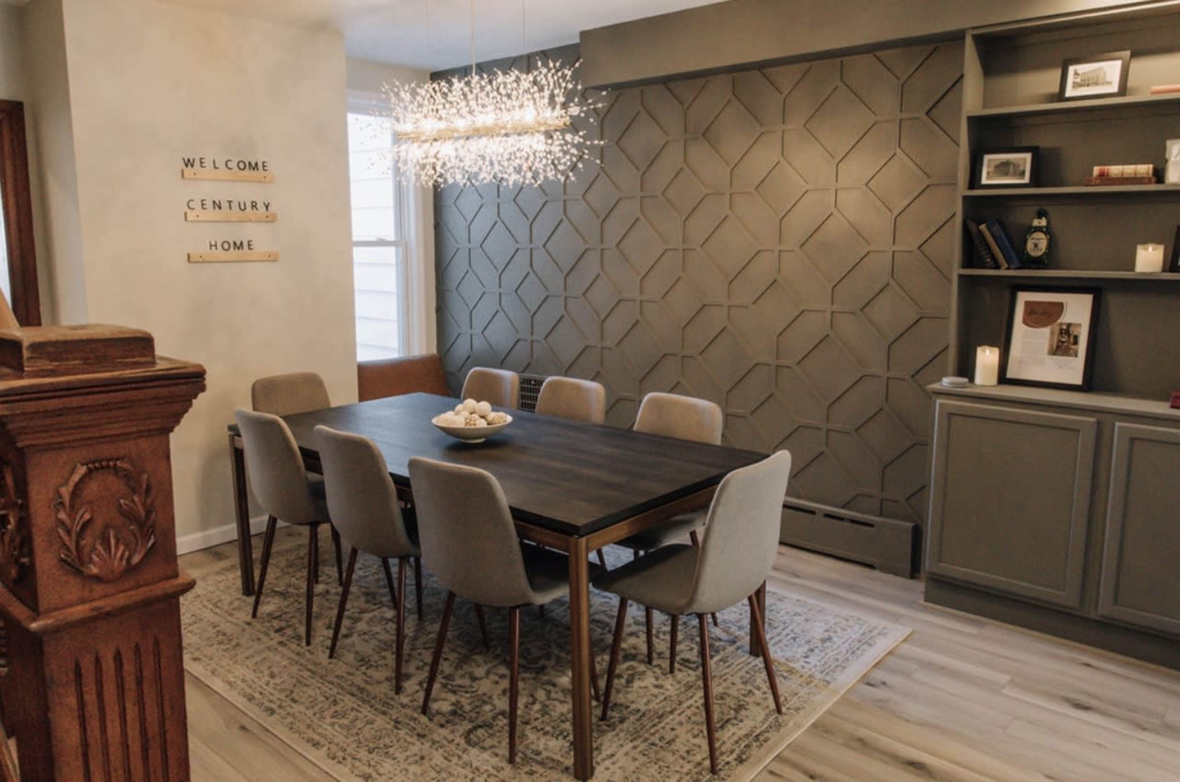 A dining room features a rectangular table surrounded by upholstered chairs, with a modern light fixture hanging above and a geometric patterned wall in the background.
