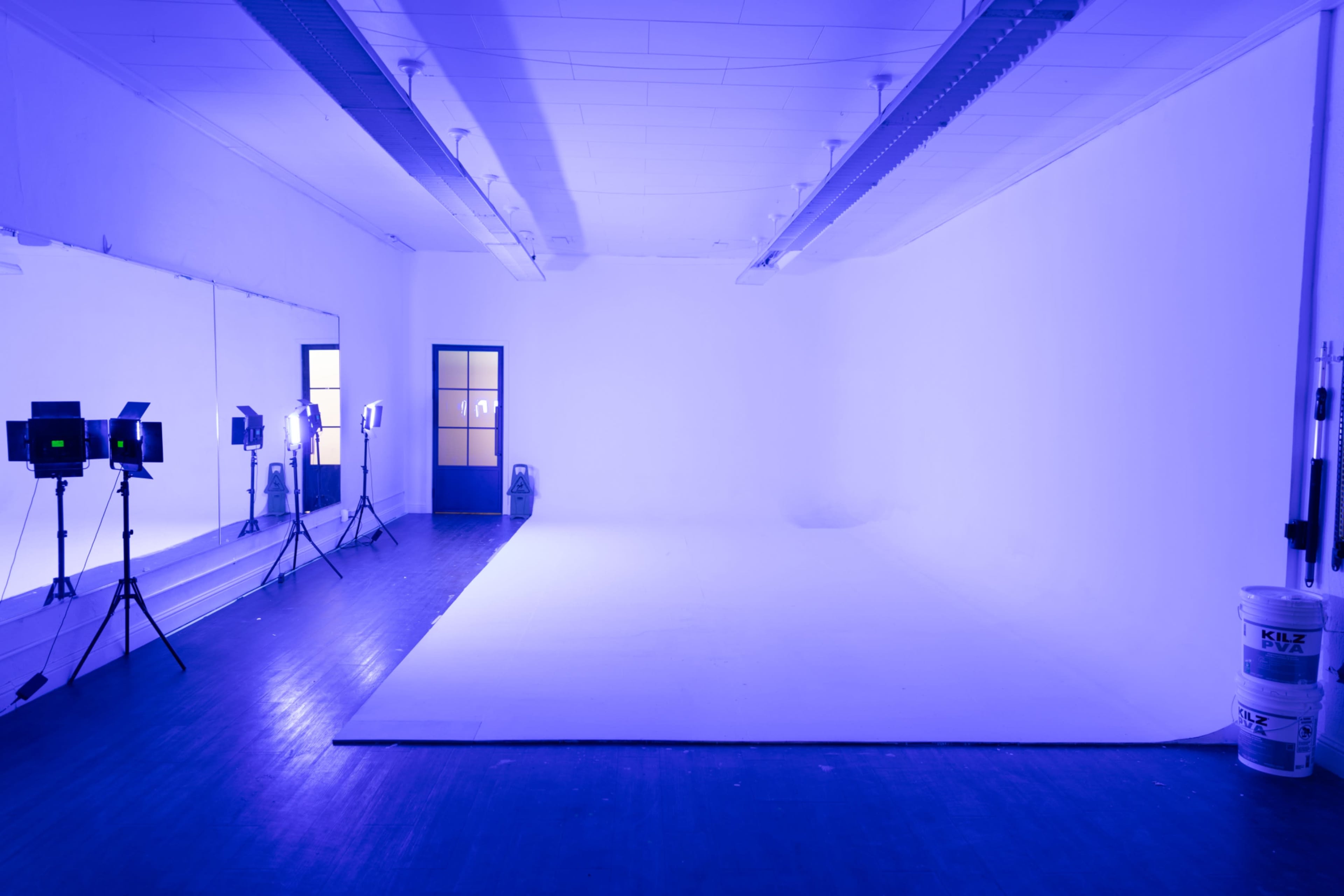 An empty studio space is illuminated with blue lighting, featuring a reflective mirror on one wall and a door with a window at the far end.