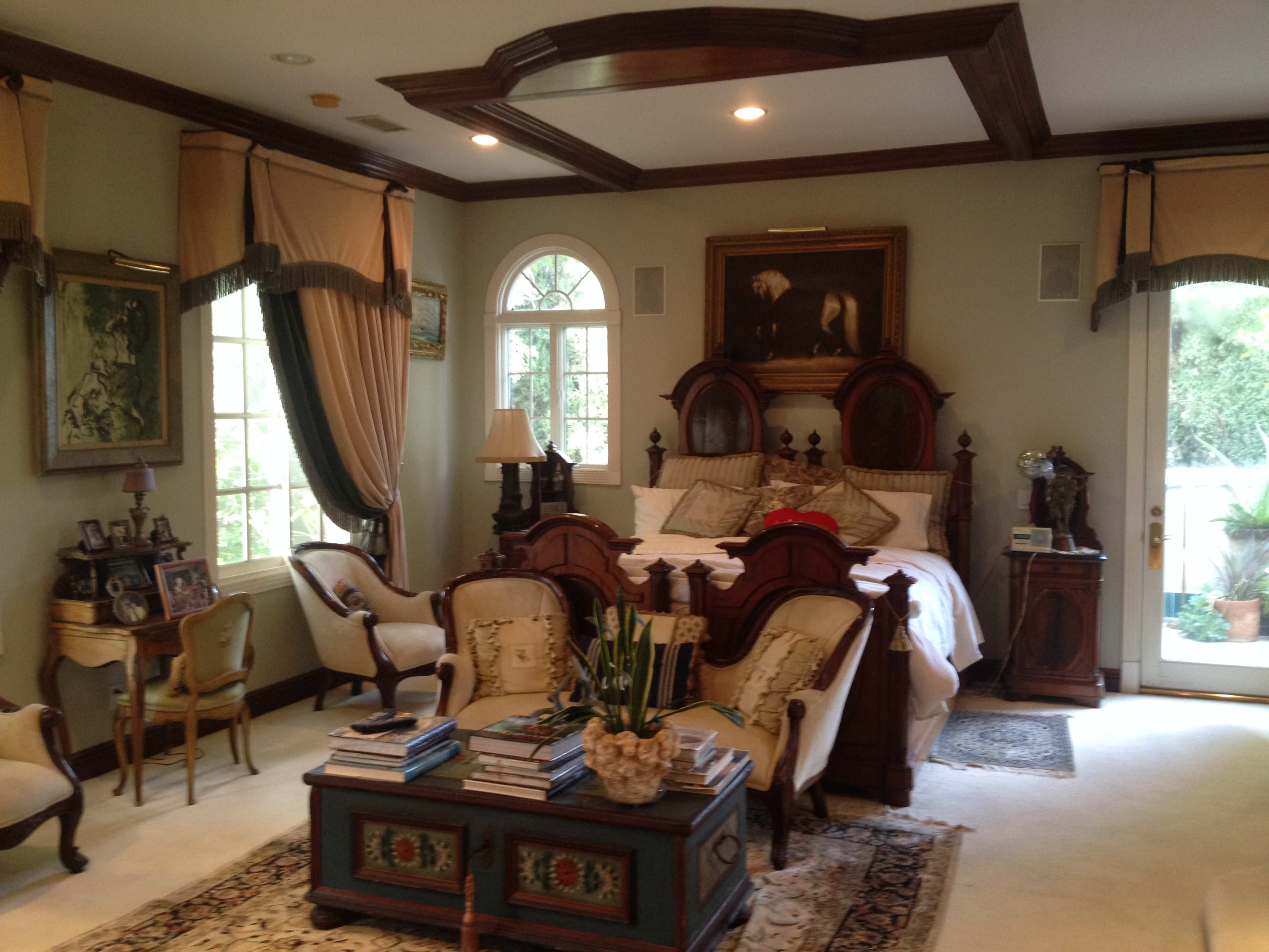 A well-furnished bedroom featuring a large wooden bed, two elegant armchairs, and a coffee table stacked with books, all arranged around a window with natural light.