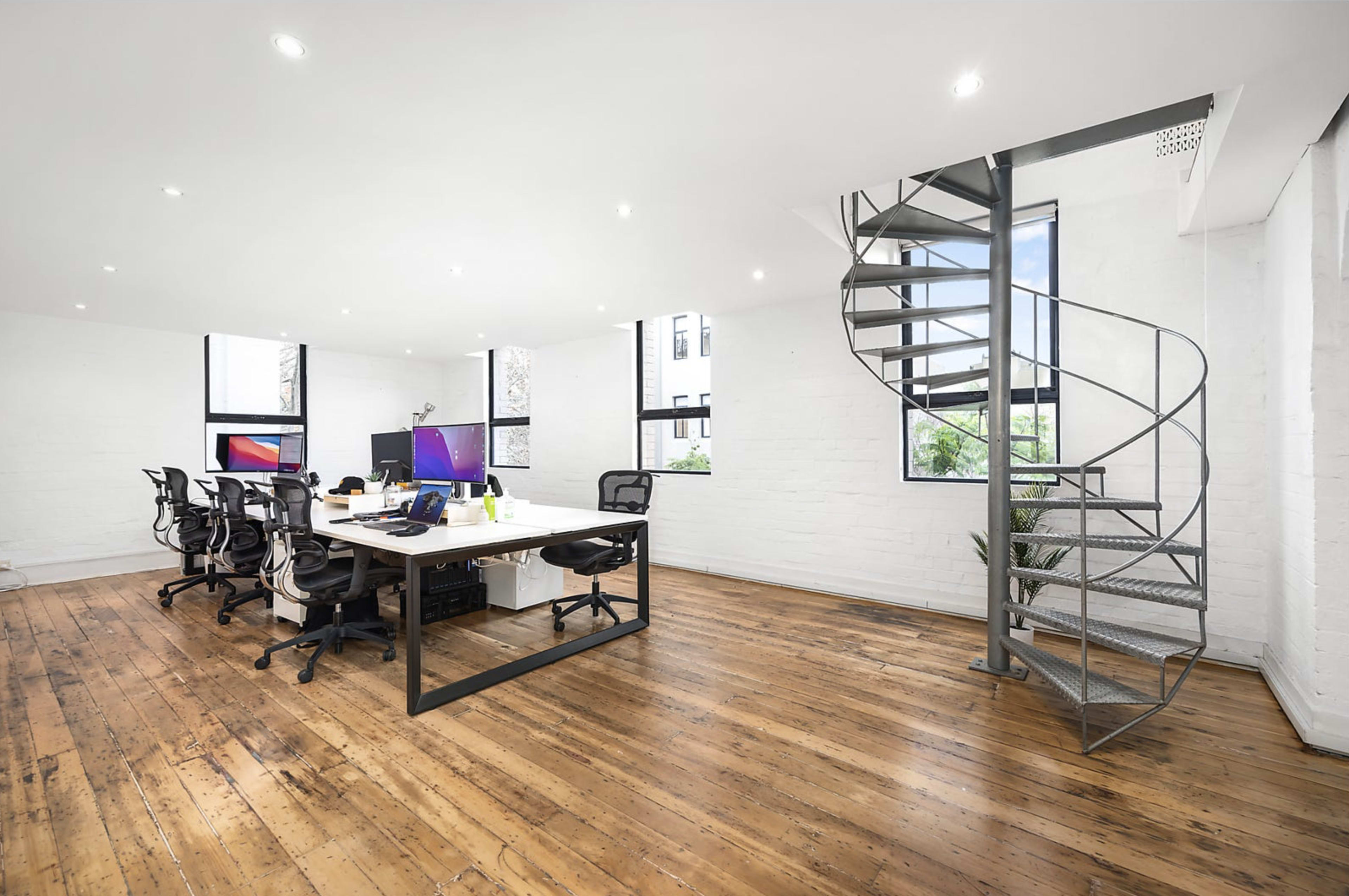 The image shows a modern office space with a large wooden floor, a long desk with multiple chairs, dual monitors, and a spiral staircase in the corner.