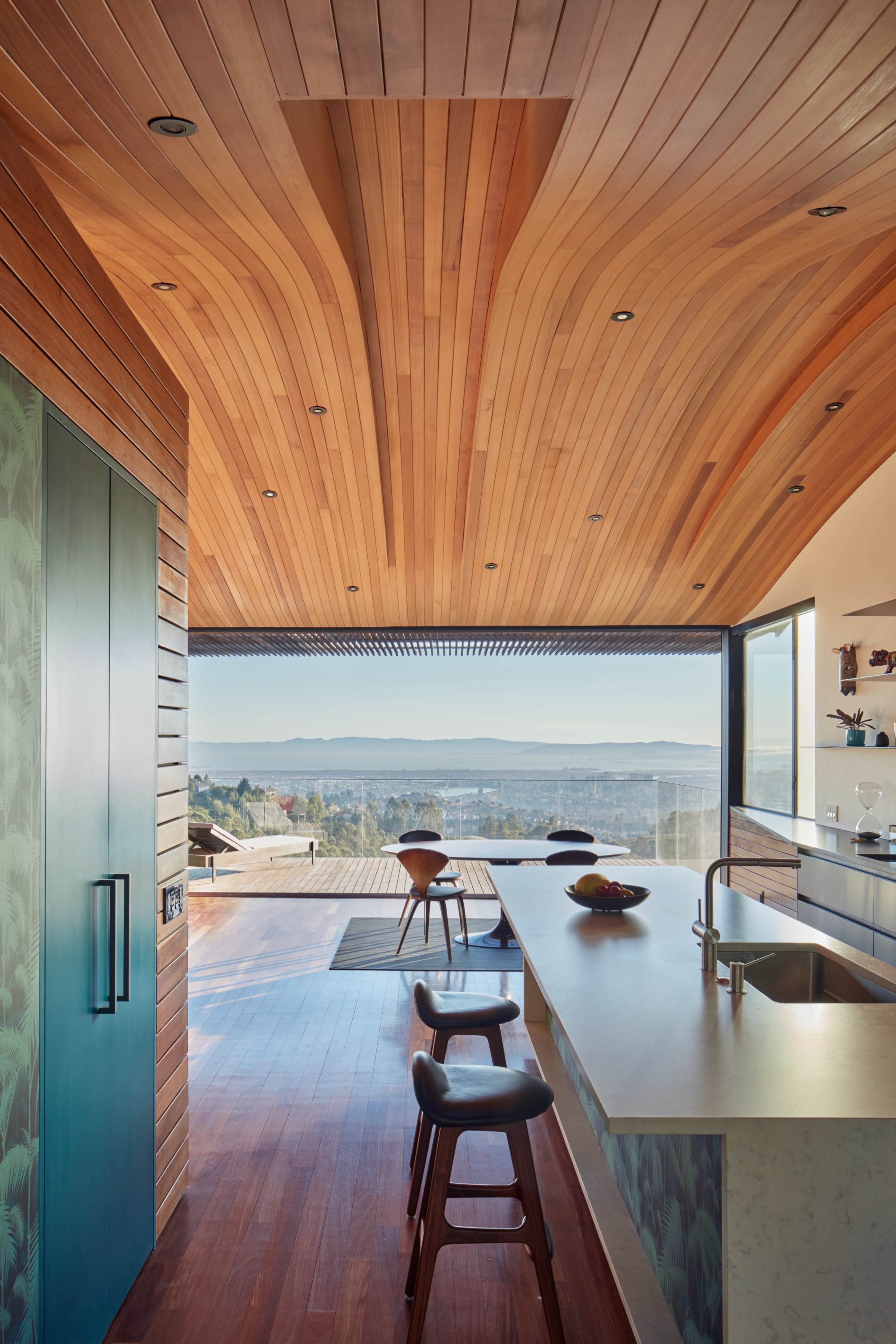 A modern kitchen features a wooden ceiling with a curved design, stainless steel appliances, and a view of a landscape through large glass doors.