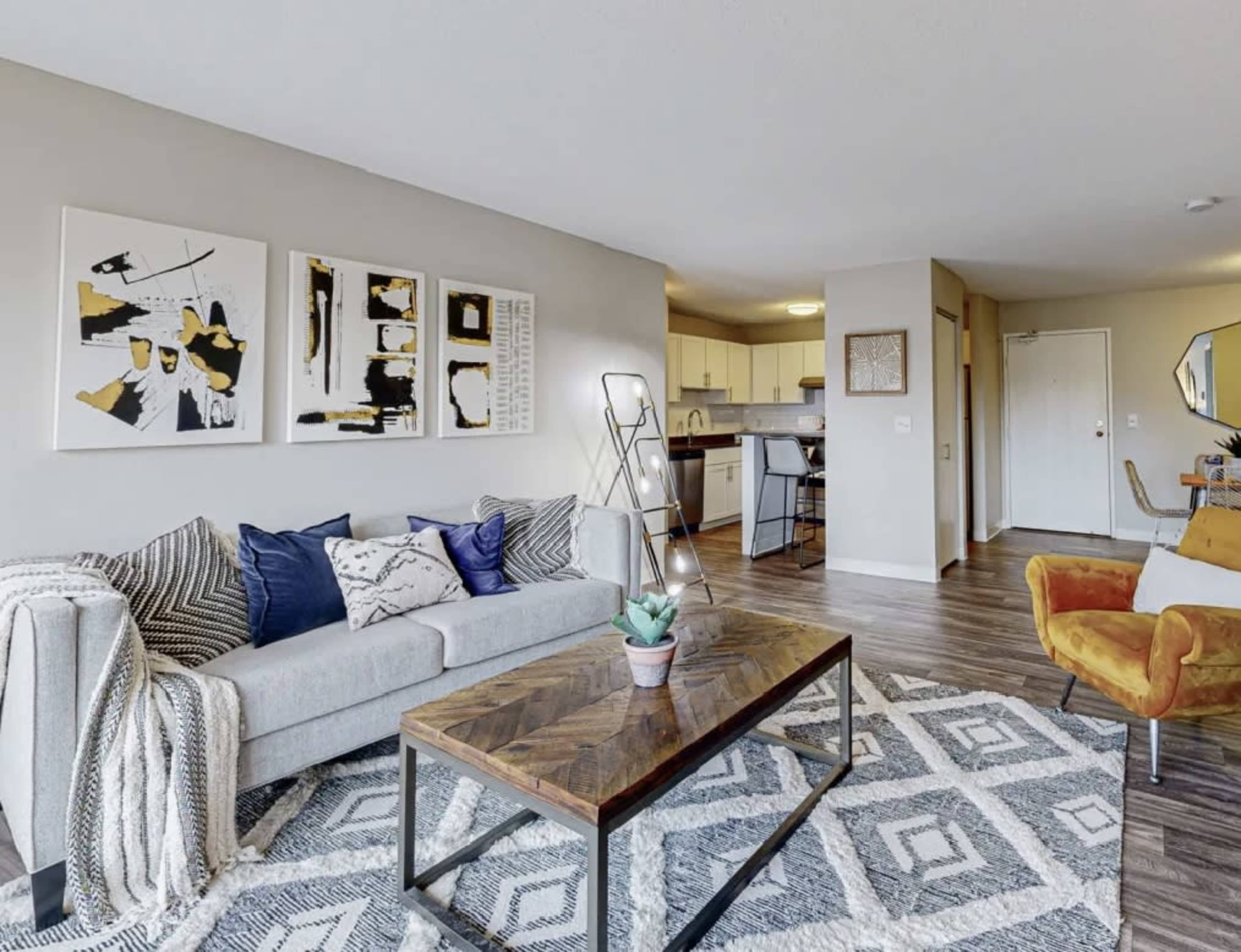 A modern living room with a sofa, decorative pillows, a coffee table, and wall art, leading into an open kitchen area.