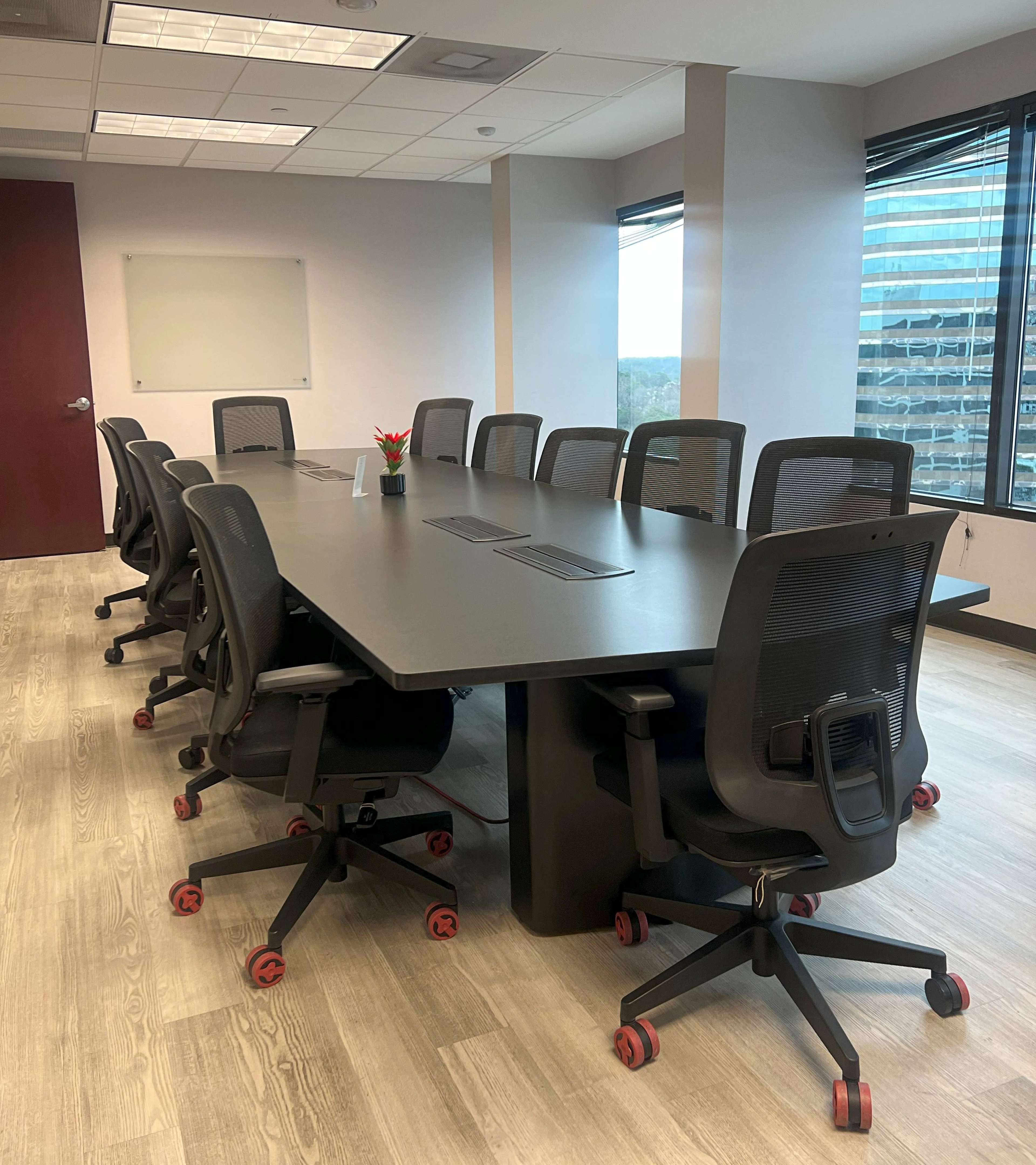 A large conference room features a long black table surrounded by ergonomic chairs on wheels, with windows displaying a cityscape in the background.