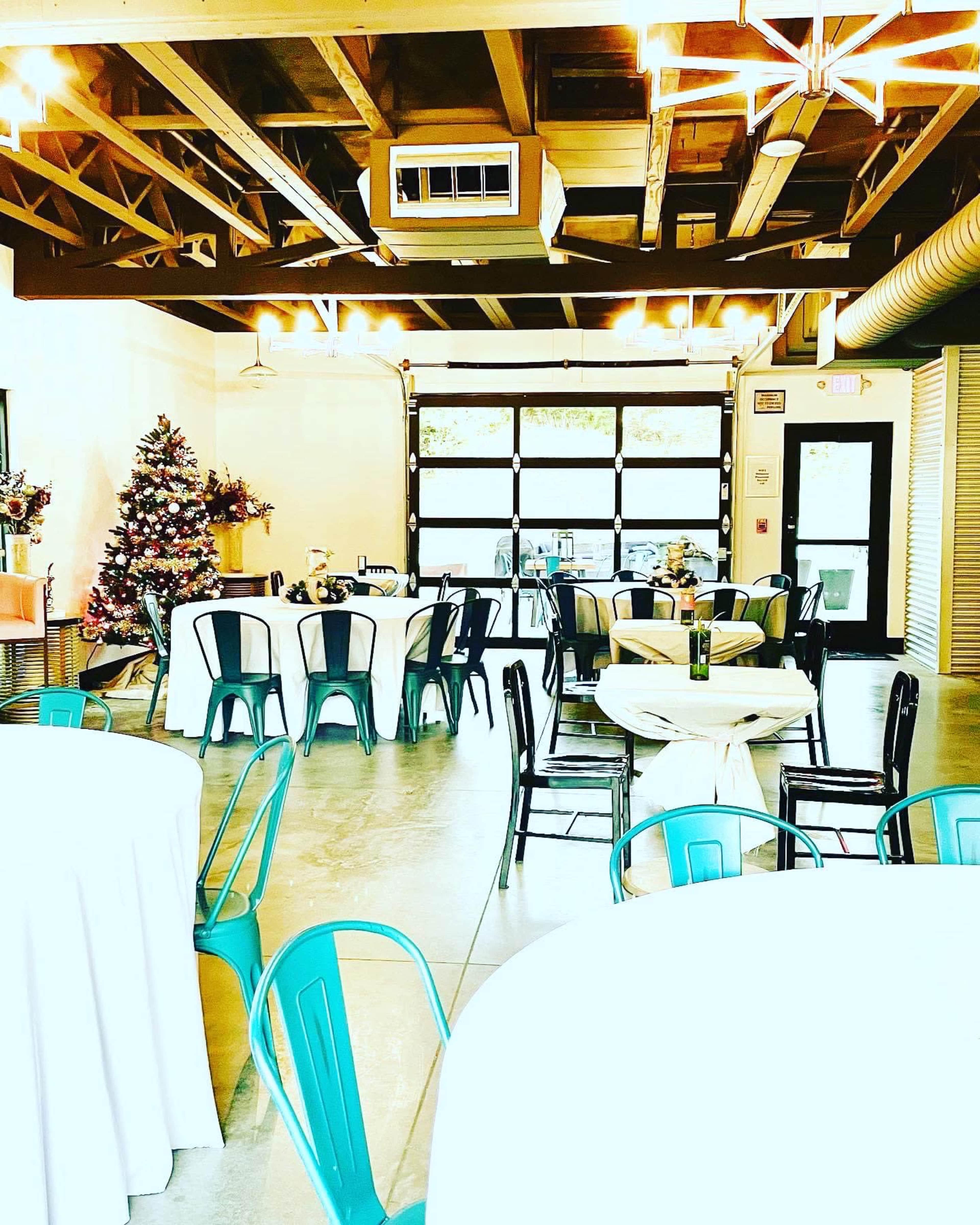 An airy event space with tables covered in white linens, metal chairs, and a decorated Christmas tree in a corner under exposed wooden beams.