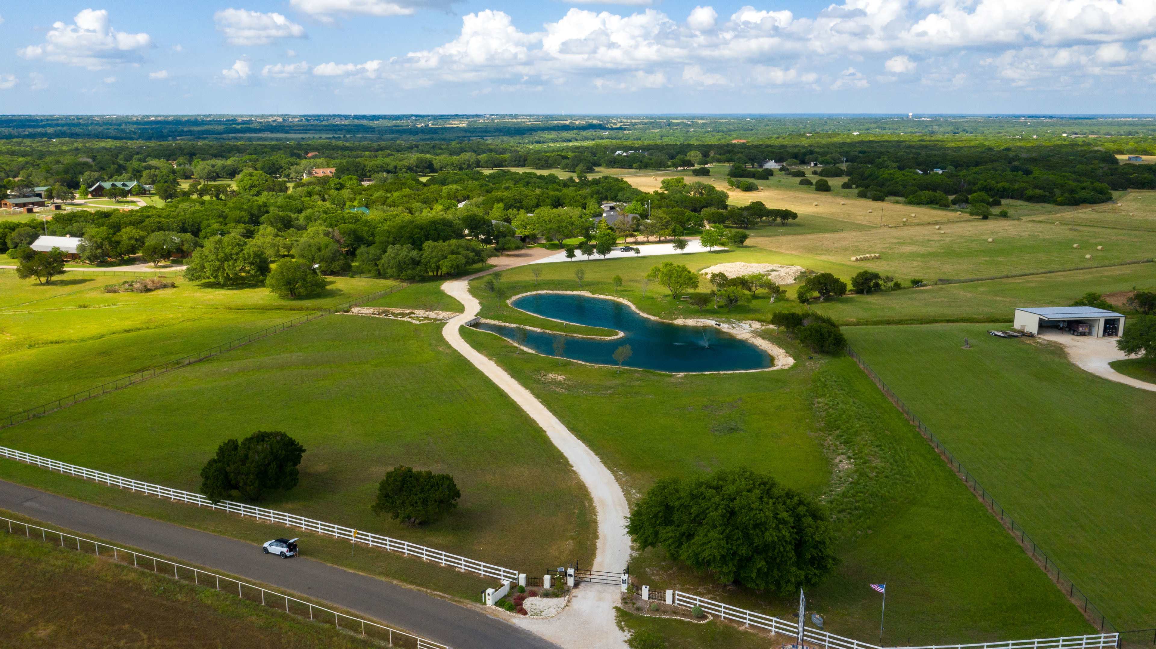 An aerial view shows a winding road leading to a pond surrounded by green fields and trees.
